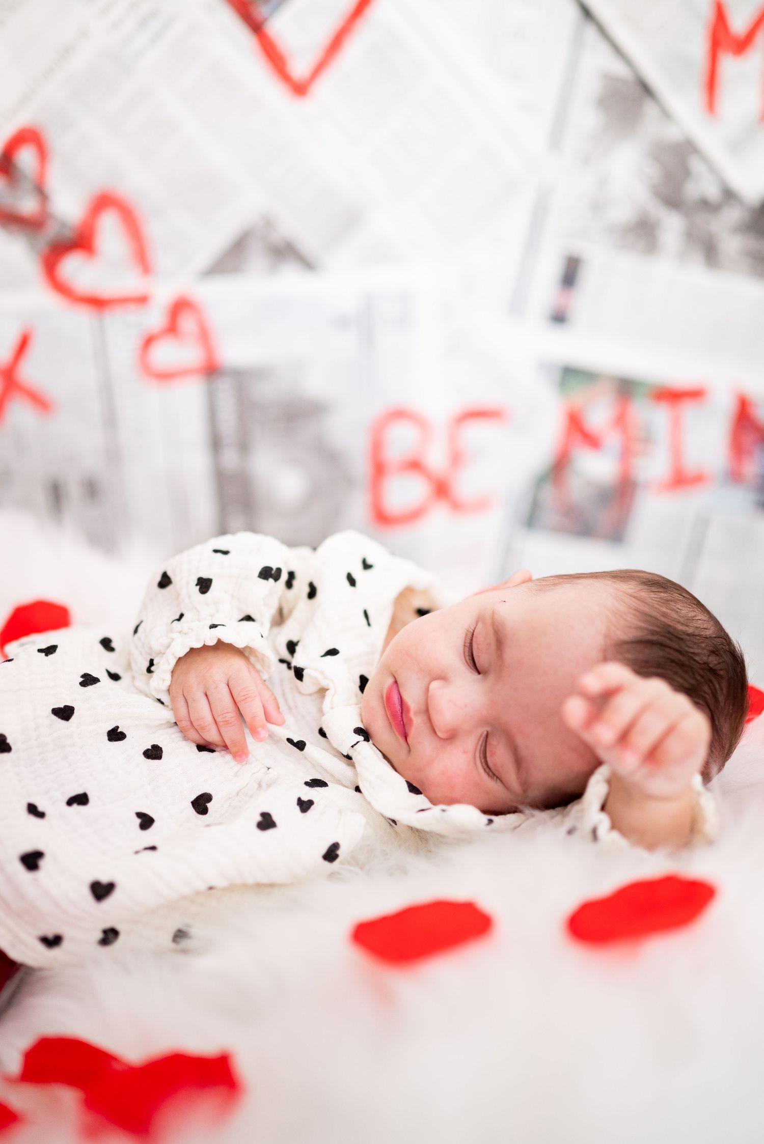 Sleeping newborn baby in heart-print onesie surrounded by red rose petals with Valentine's Day decorations.