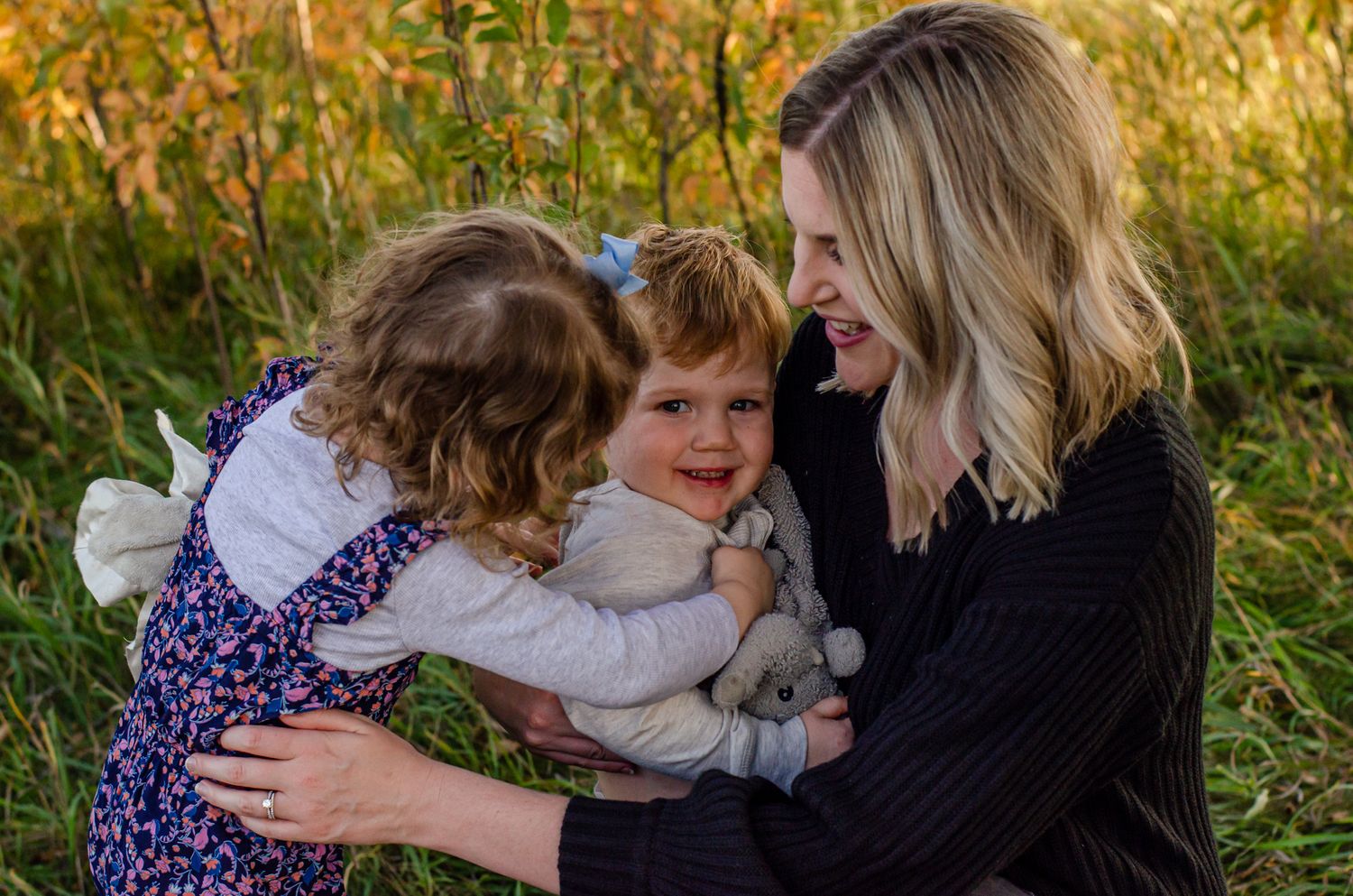 A smiling blonde woman hugs two young curly-haired girls in a sunlit grassy field during a family photo session.