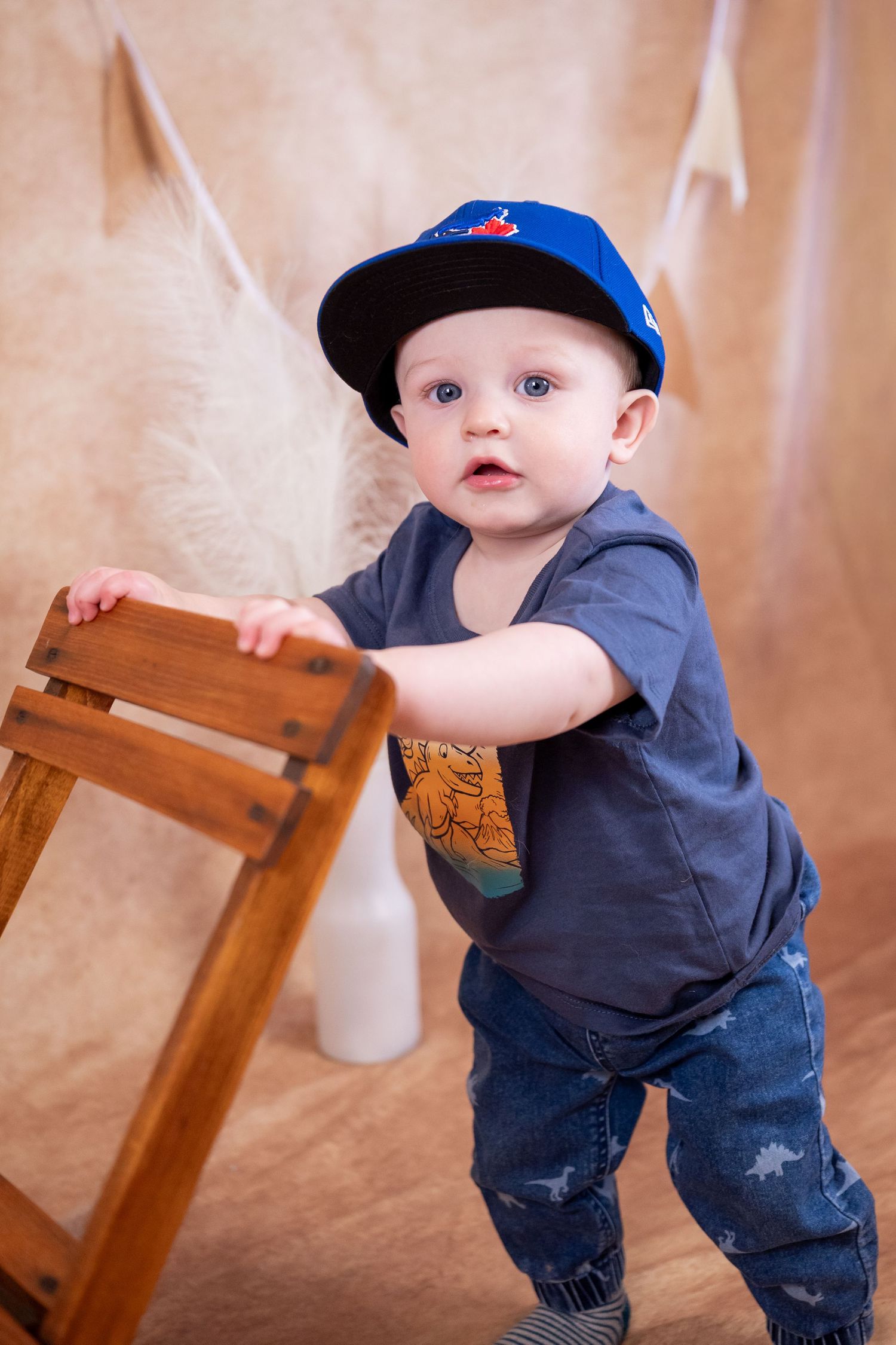 Adorable baby boy in blue cap and jeans stands holding wooden ladder in studio photo session.