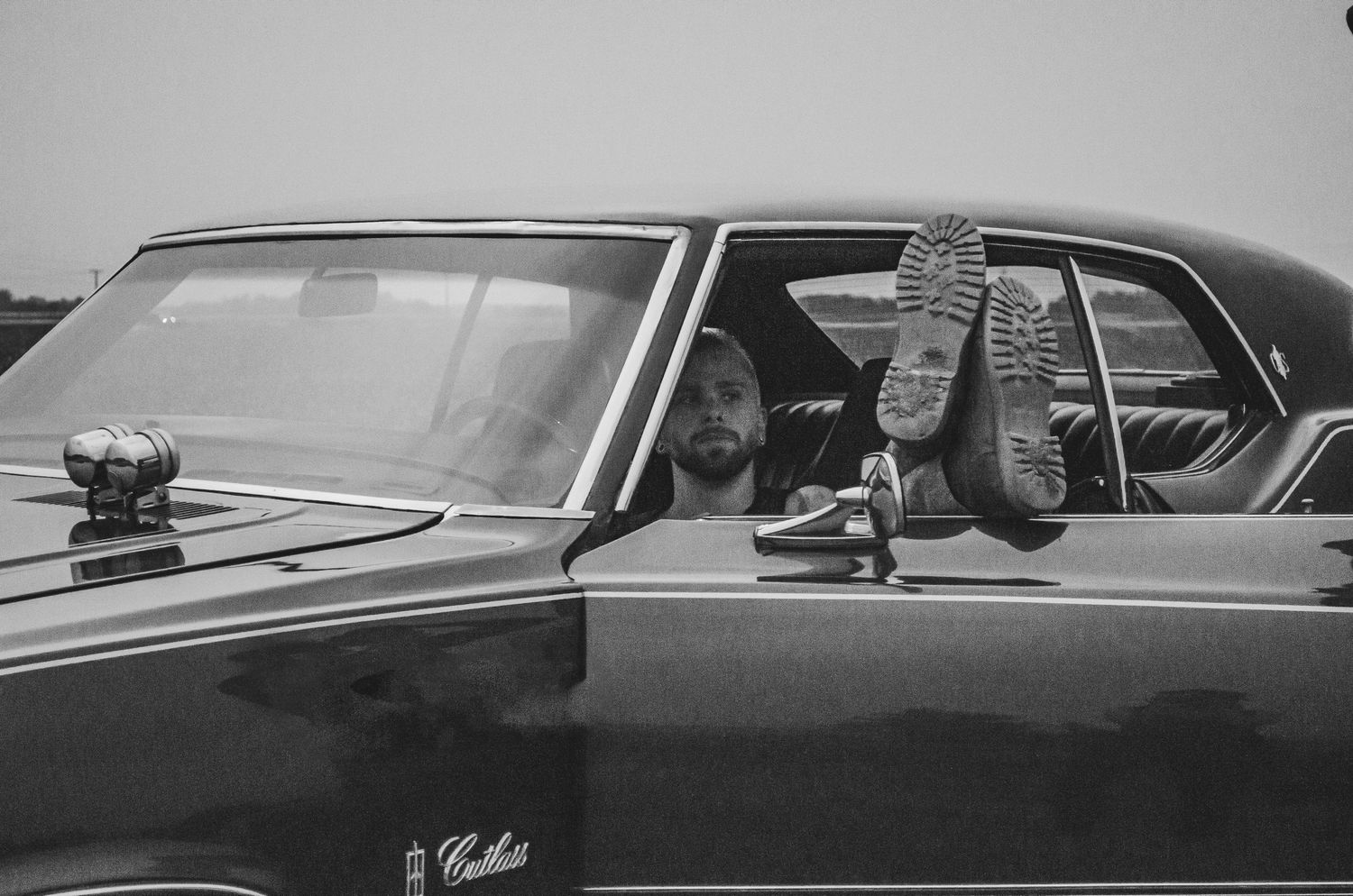 Black and white photo of two people relaxing in a classic Cadillac car, one resting feet on the window.