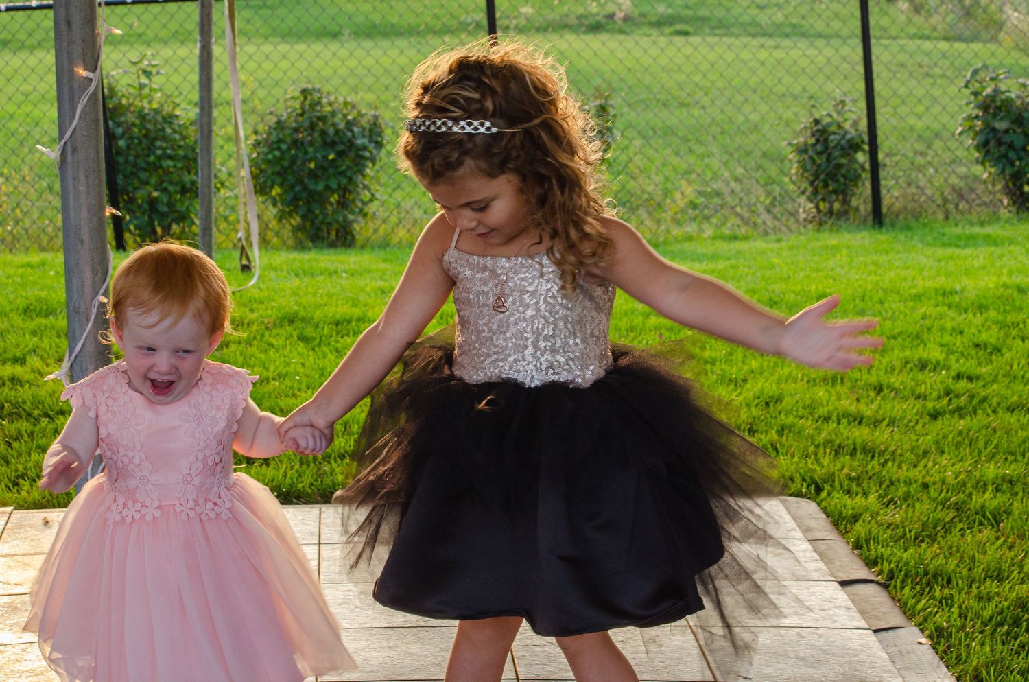 Two young girls in elegant dresses dance outdoors, one in pink tutu and one in black and gold sequin dress.