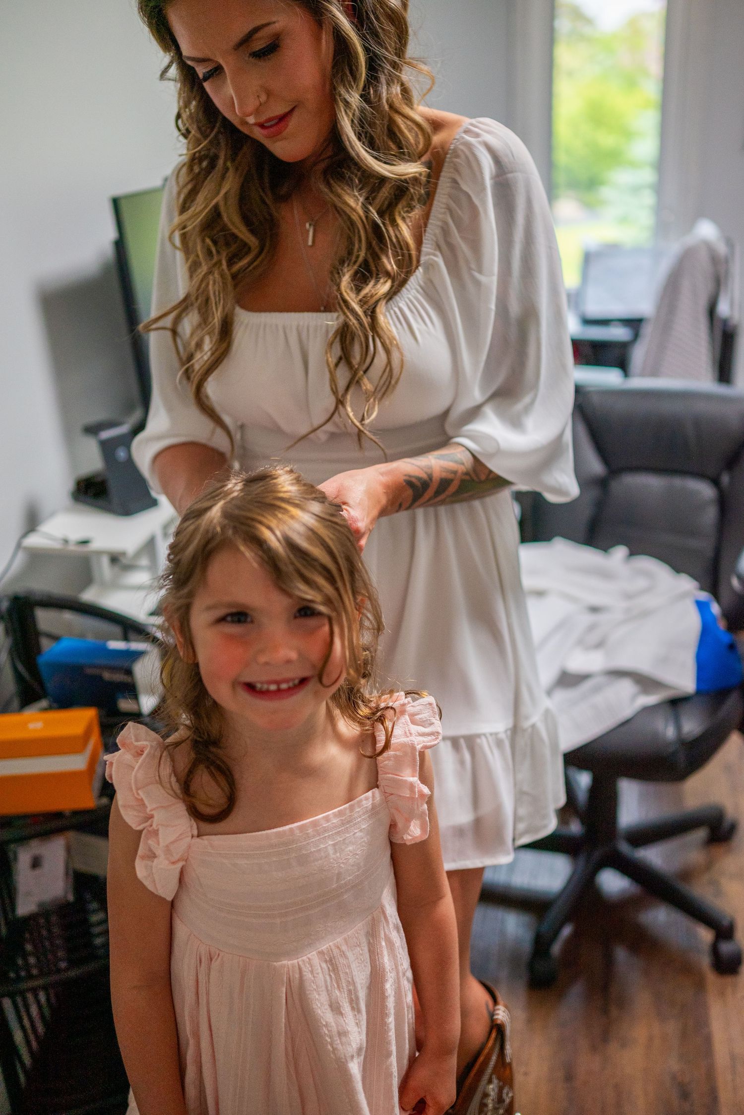 Woman in white dress styling young girl's hair, both smiling in bright room.