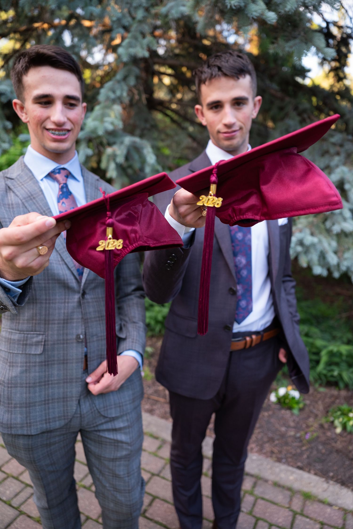 Two young men in suits holding maroon 2026 graduation caps outdoors on a brick path.