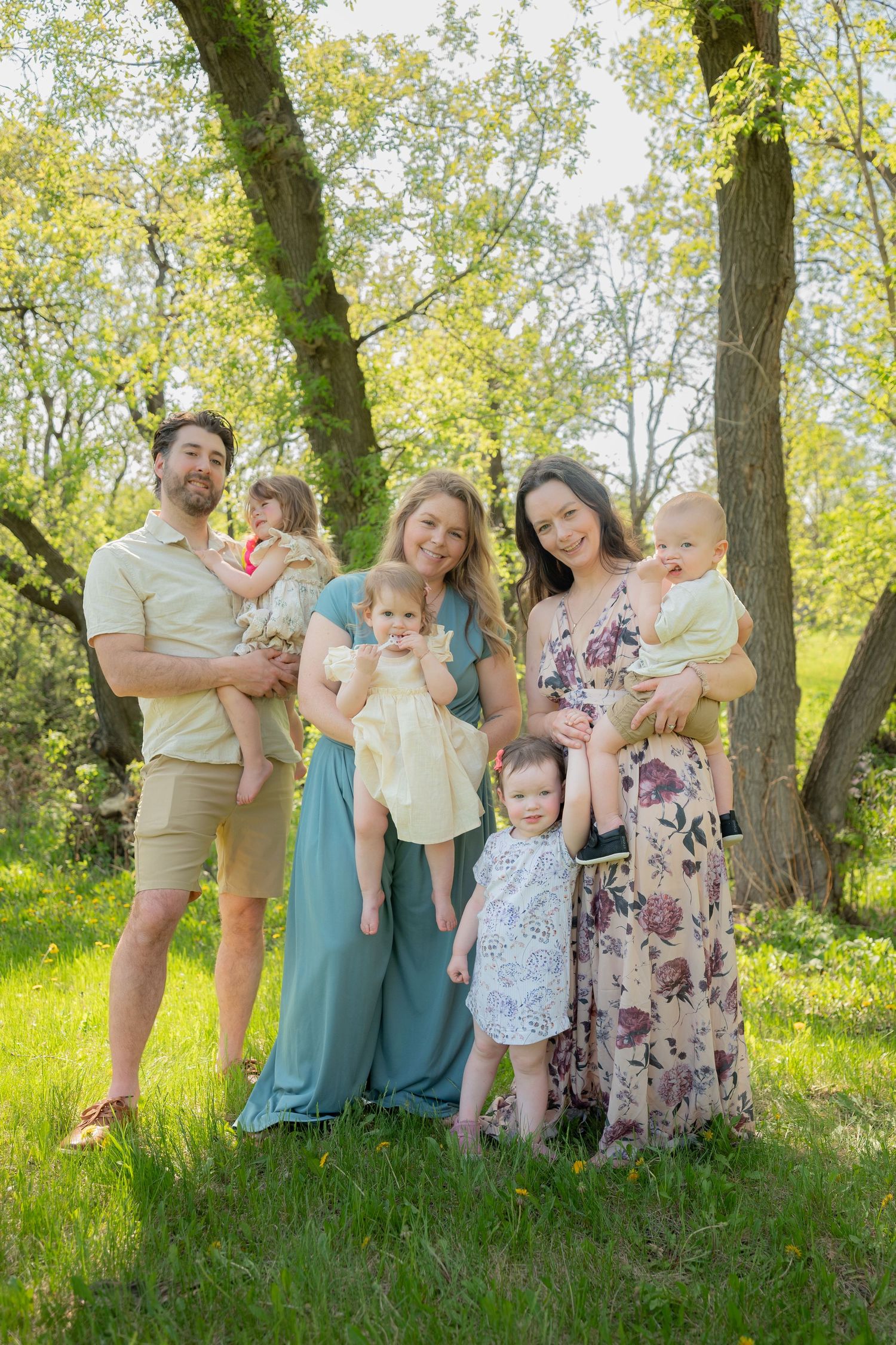 A joyful family of two adults and four young children posing outdoors in a lush green park on a sunny day.
