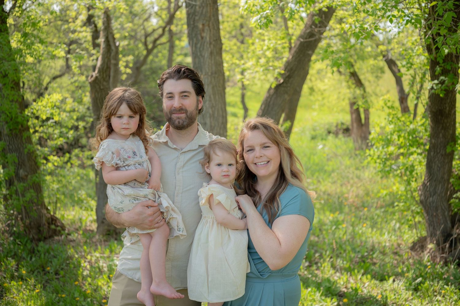 Family of four smiling outdoors in lush green forest, parents holding two young daughters in spring nature setting.