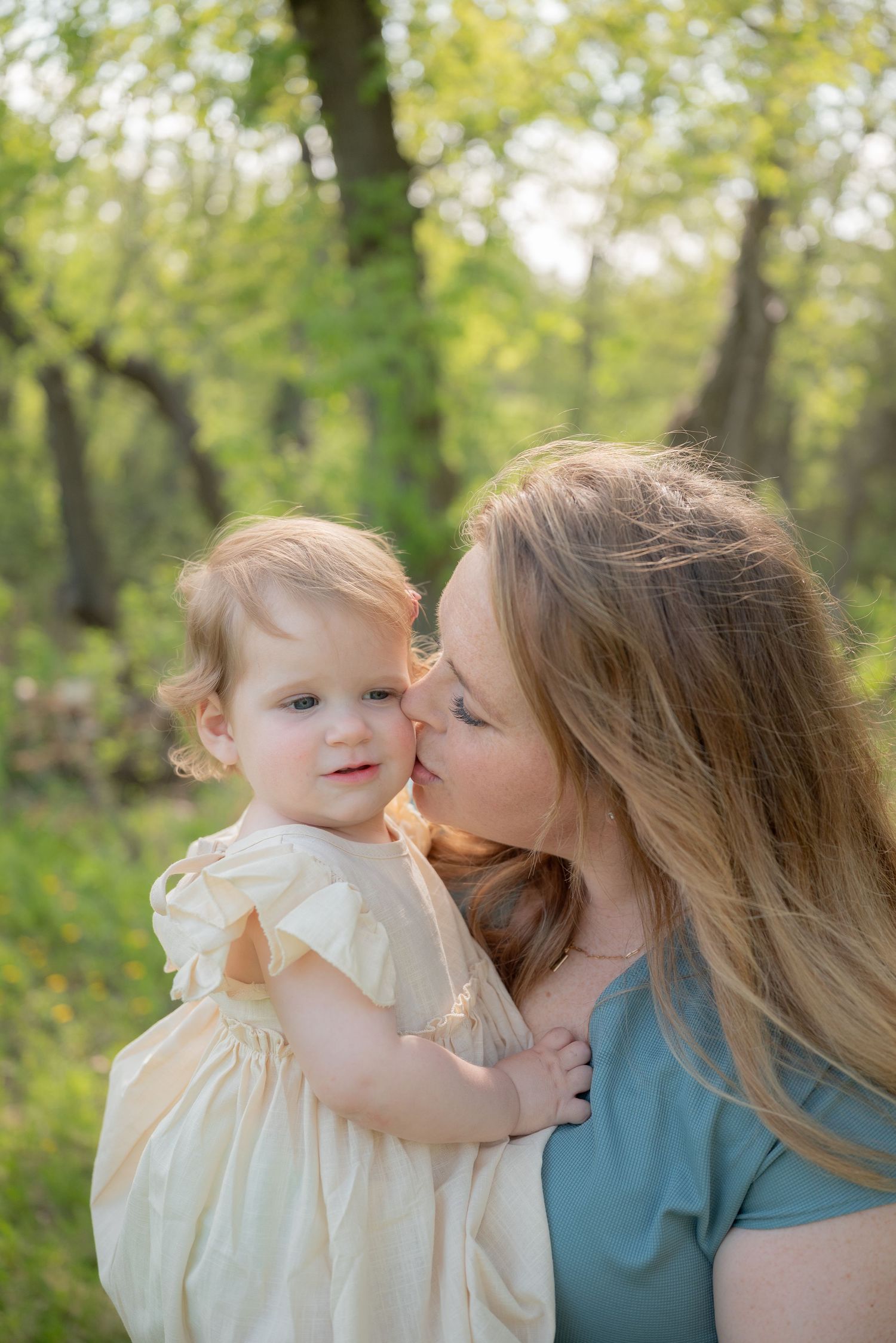 A mother tenderly kisses her toddler daughter outdoors, both in light clothing, surrounded by lush green trees.