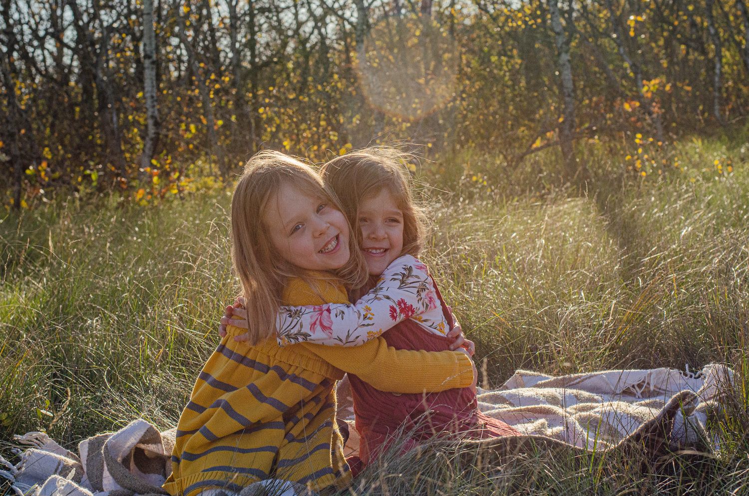 Two young blonde girls hugging and smiling outdoors on a sunny day in a grassy field.