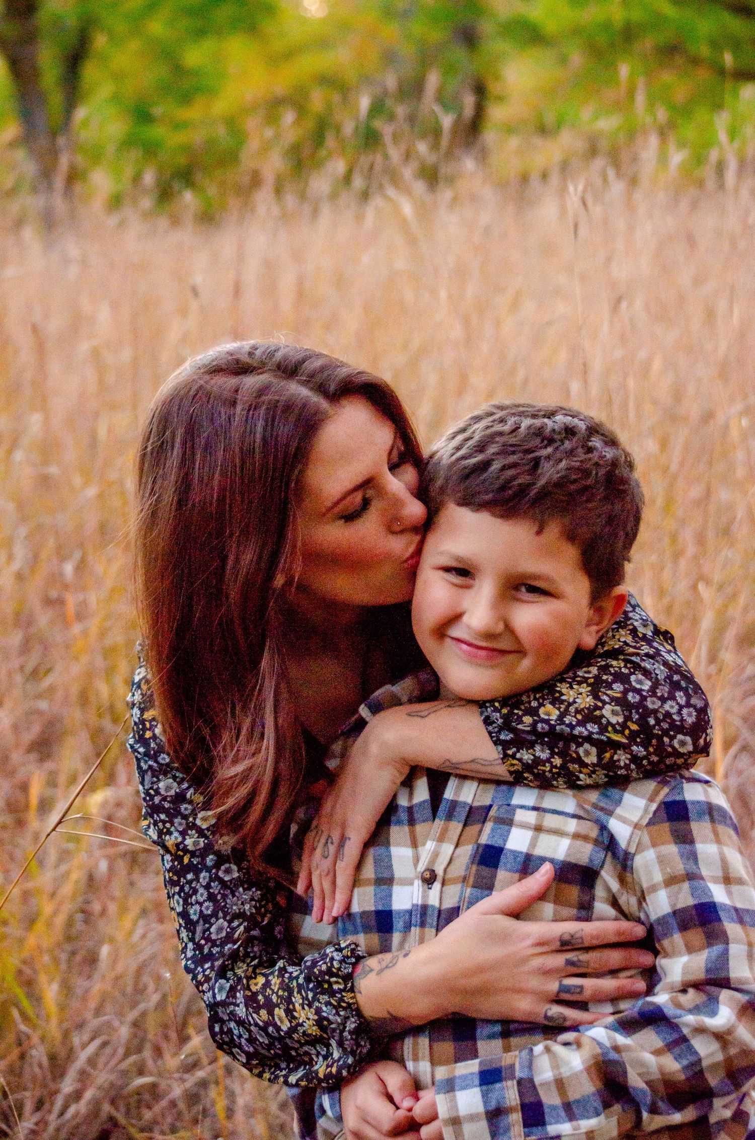 A mother hugs and kisses her smiling young son in a golden field during an outdoor fall family photo session.