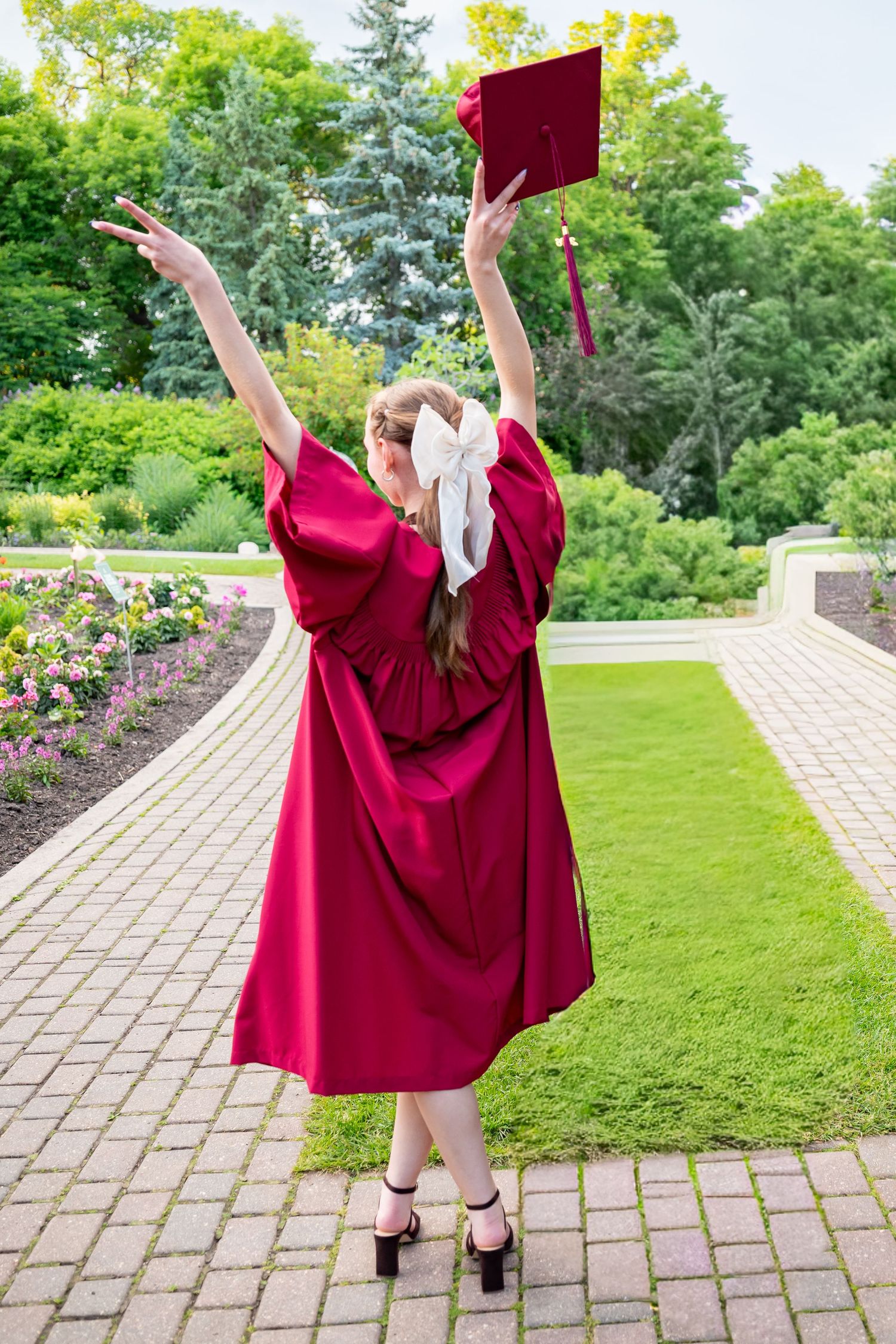 Graduate in red gown raises cap triumphantly in lush garden, celebrating academic achievement outdoors.