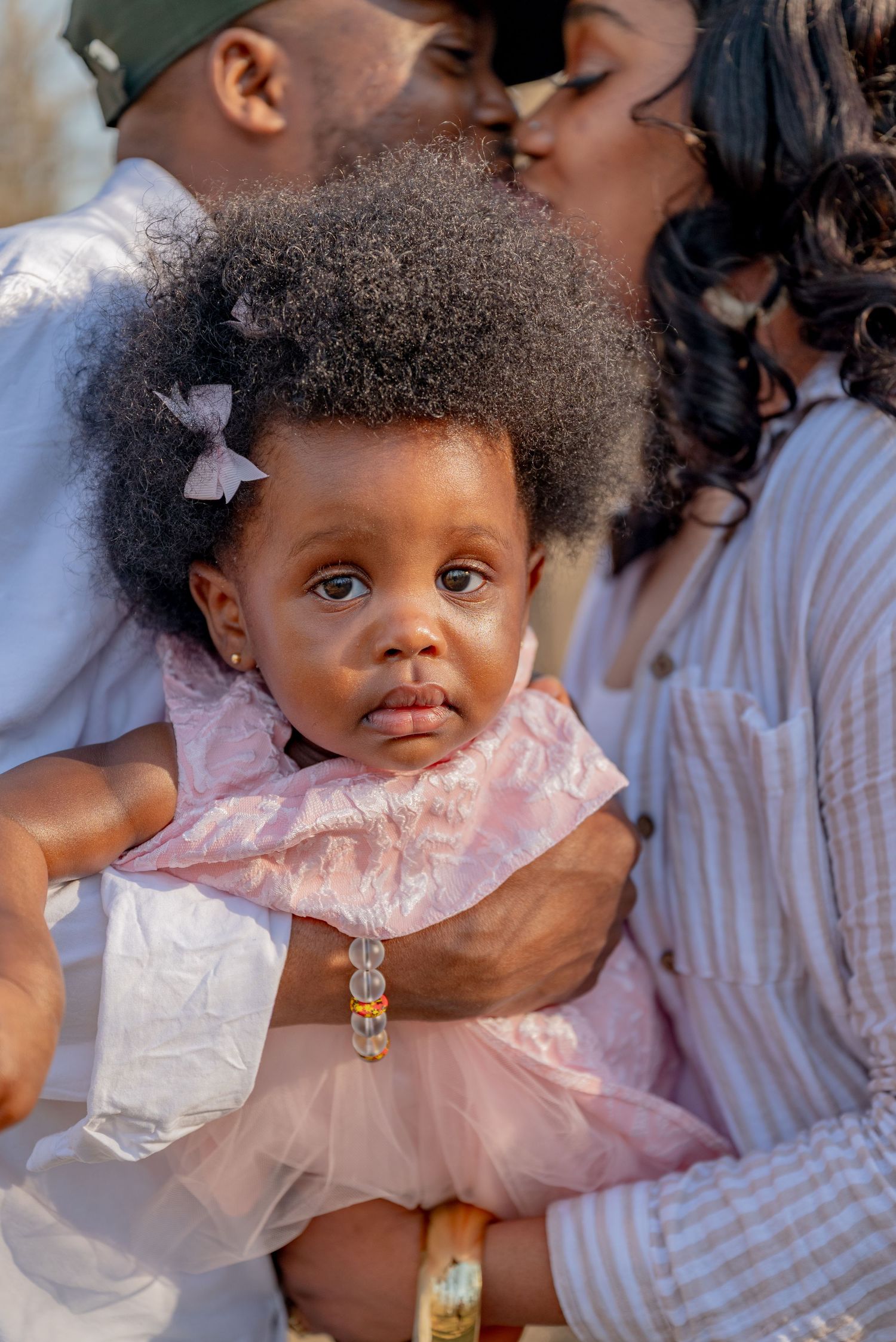 Adorable baby girl in pink dress held by parents sharing a kiss in a heartwarming family portrait.