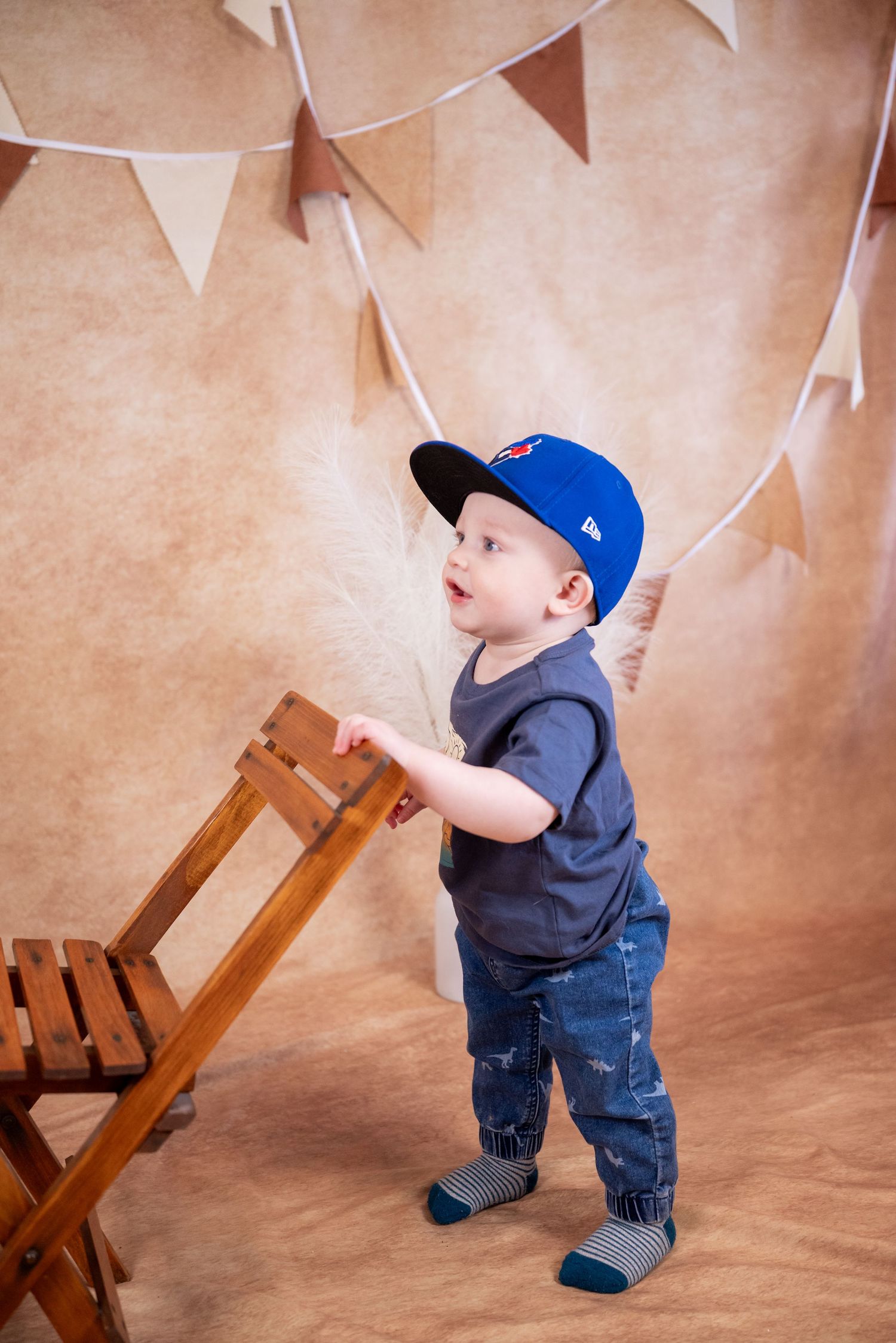 Toddler boy in blue cap and jeans standing by wooden chair with bunting backdrop.