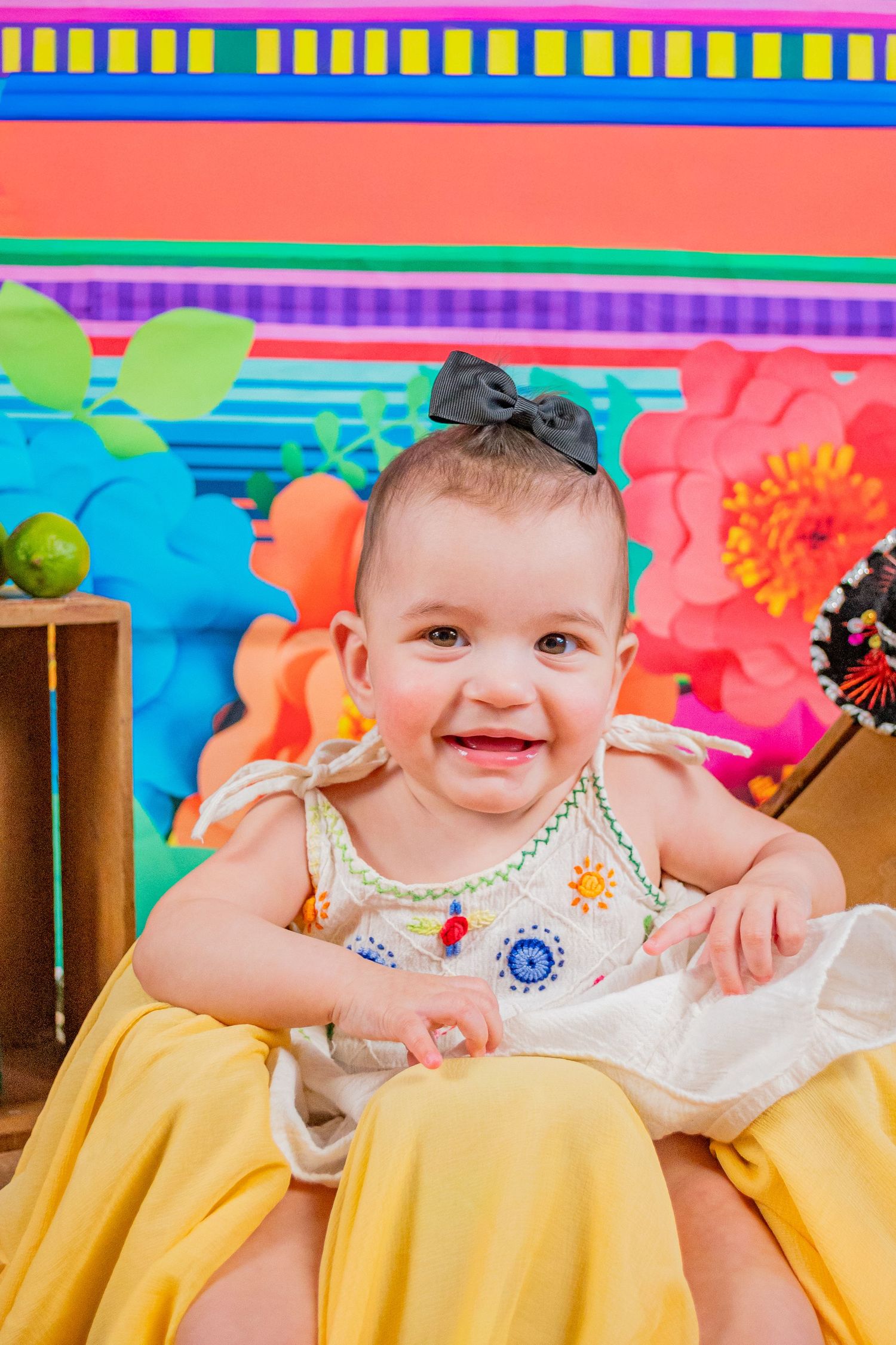 Smiling baby girl in embroidered dress with bow headband sits before colorful floral backdrop at festive party.