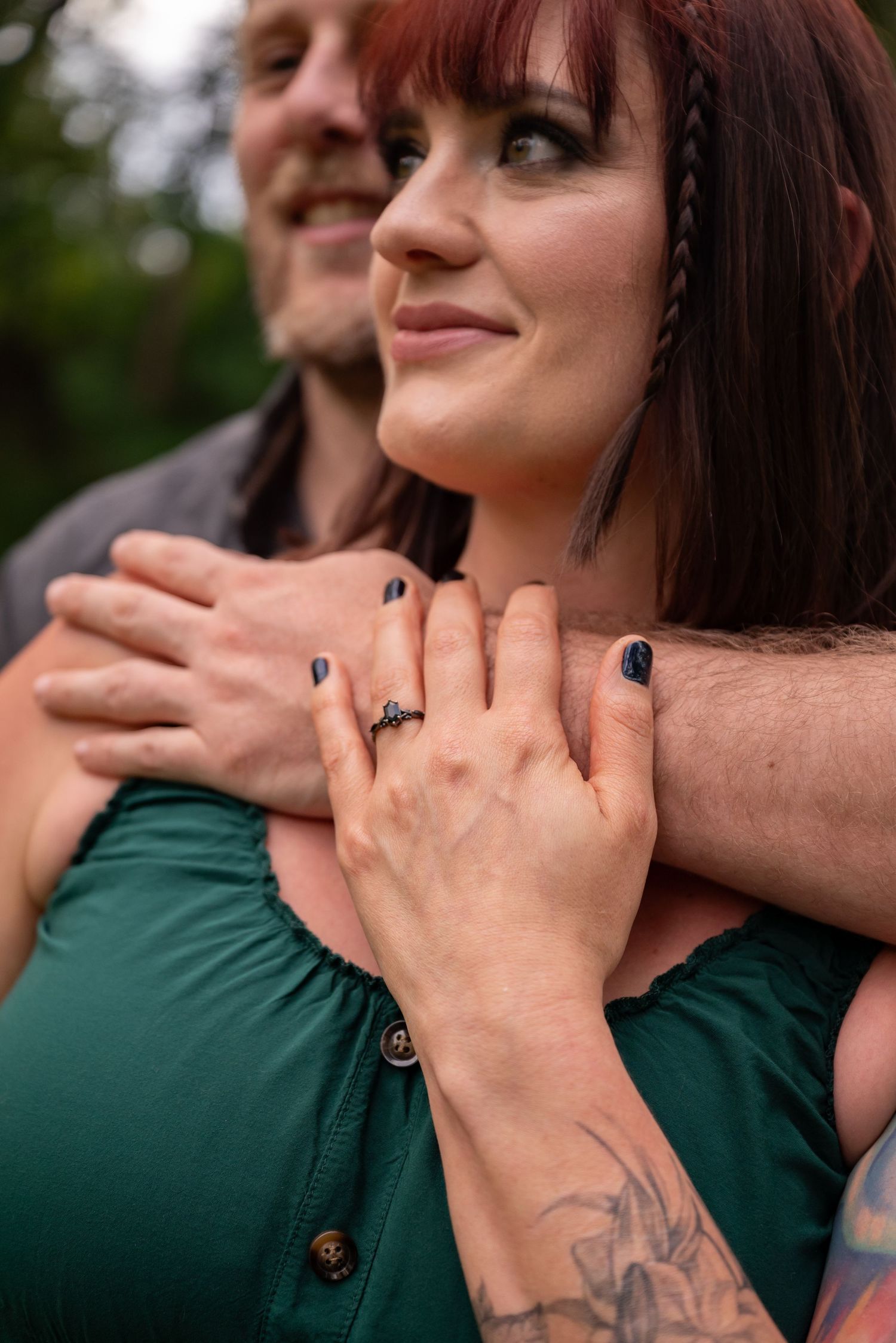Couple embracing outdoors, woman in green dress with engagement ring and dark nails, man with tattoo hugging her from behind.