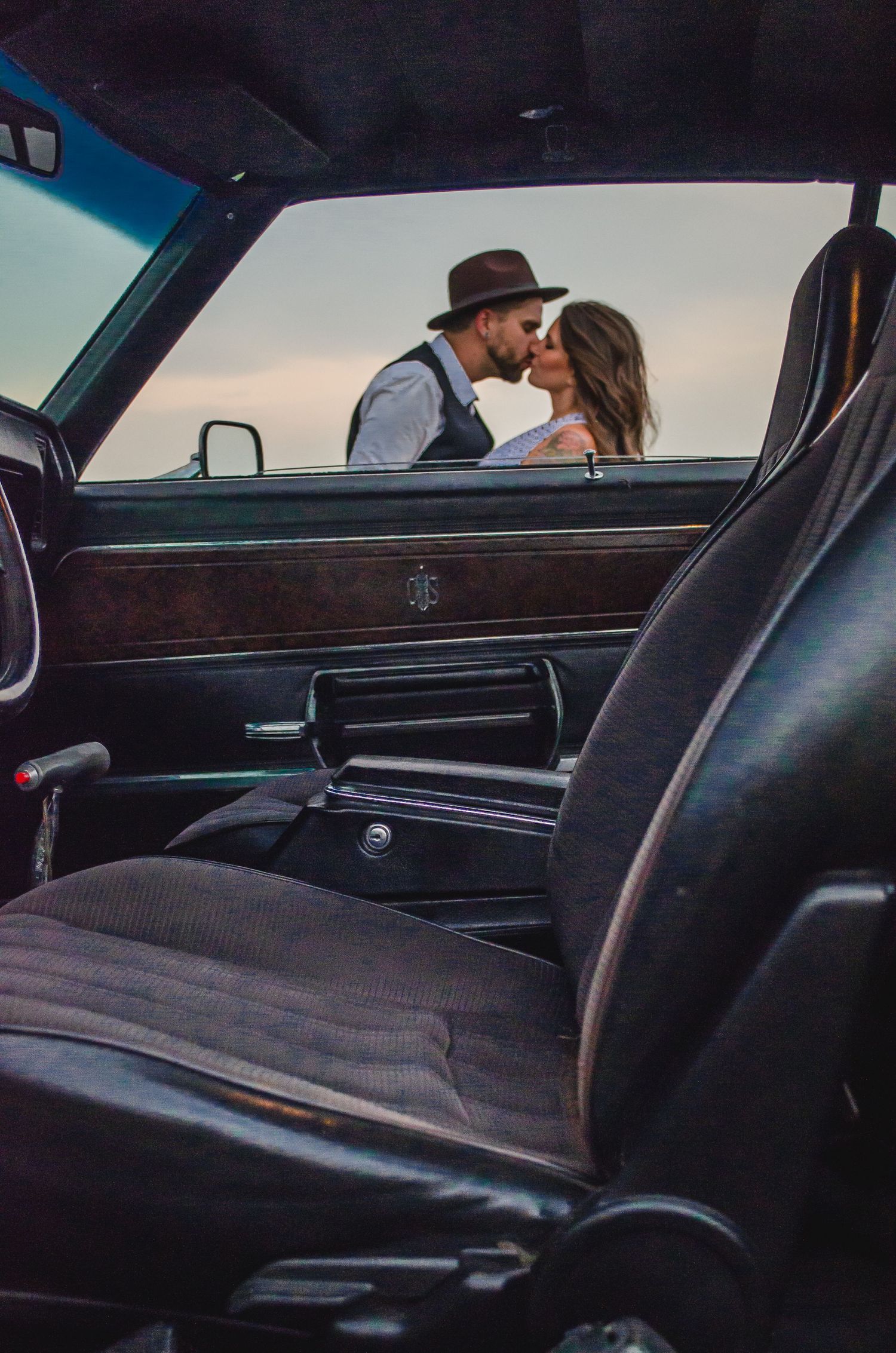 Romantic couple kissing beside a vintage classic car interior, man wearing a brown fedora hat at sunset.