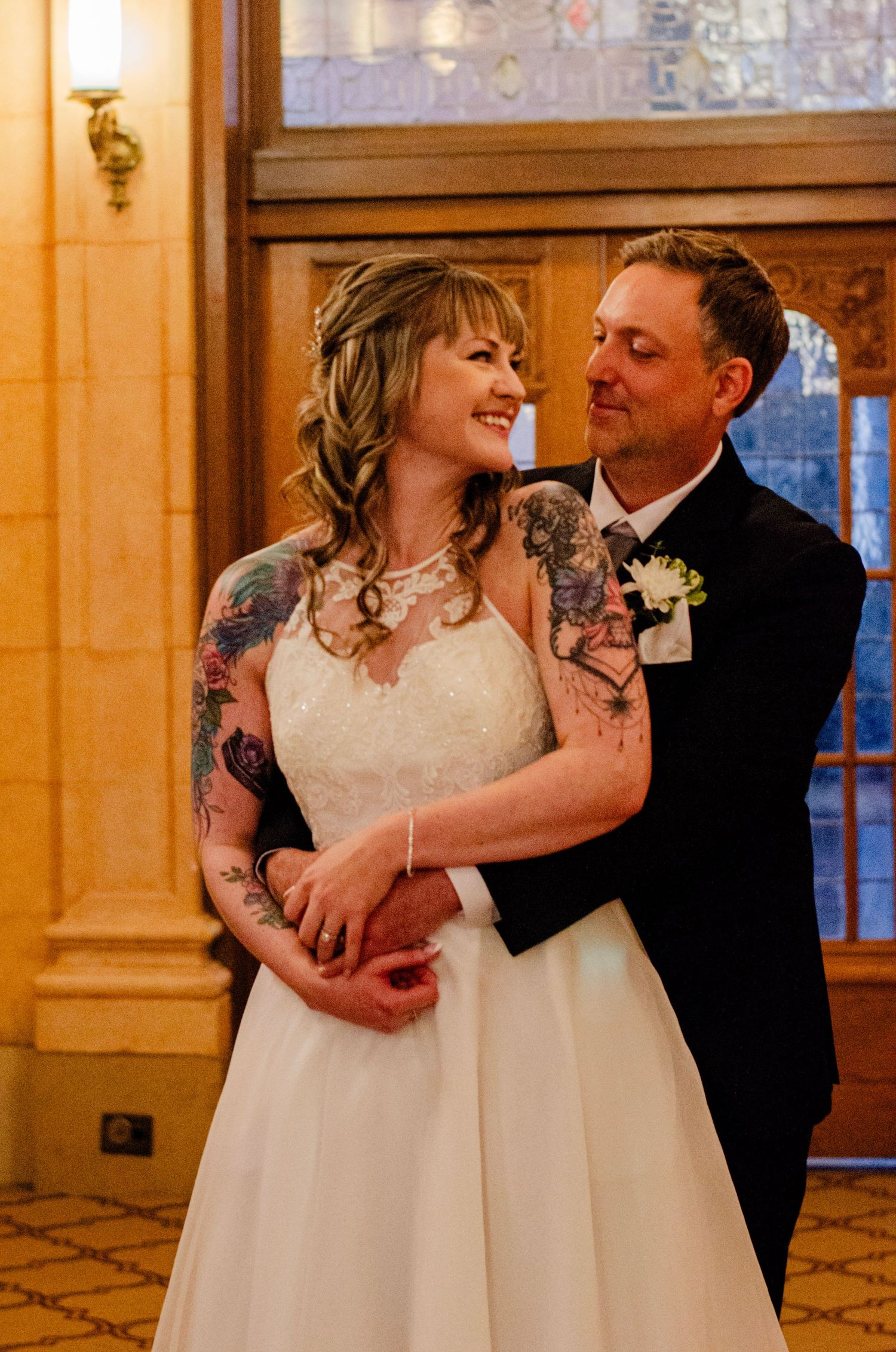 Bride with tattoos in white gown embraces groom in navy suit, sharing a tender moment in an elegant venue.