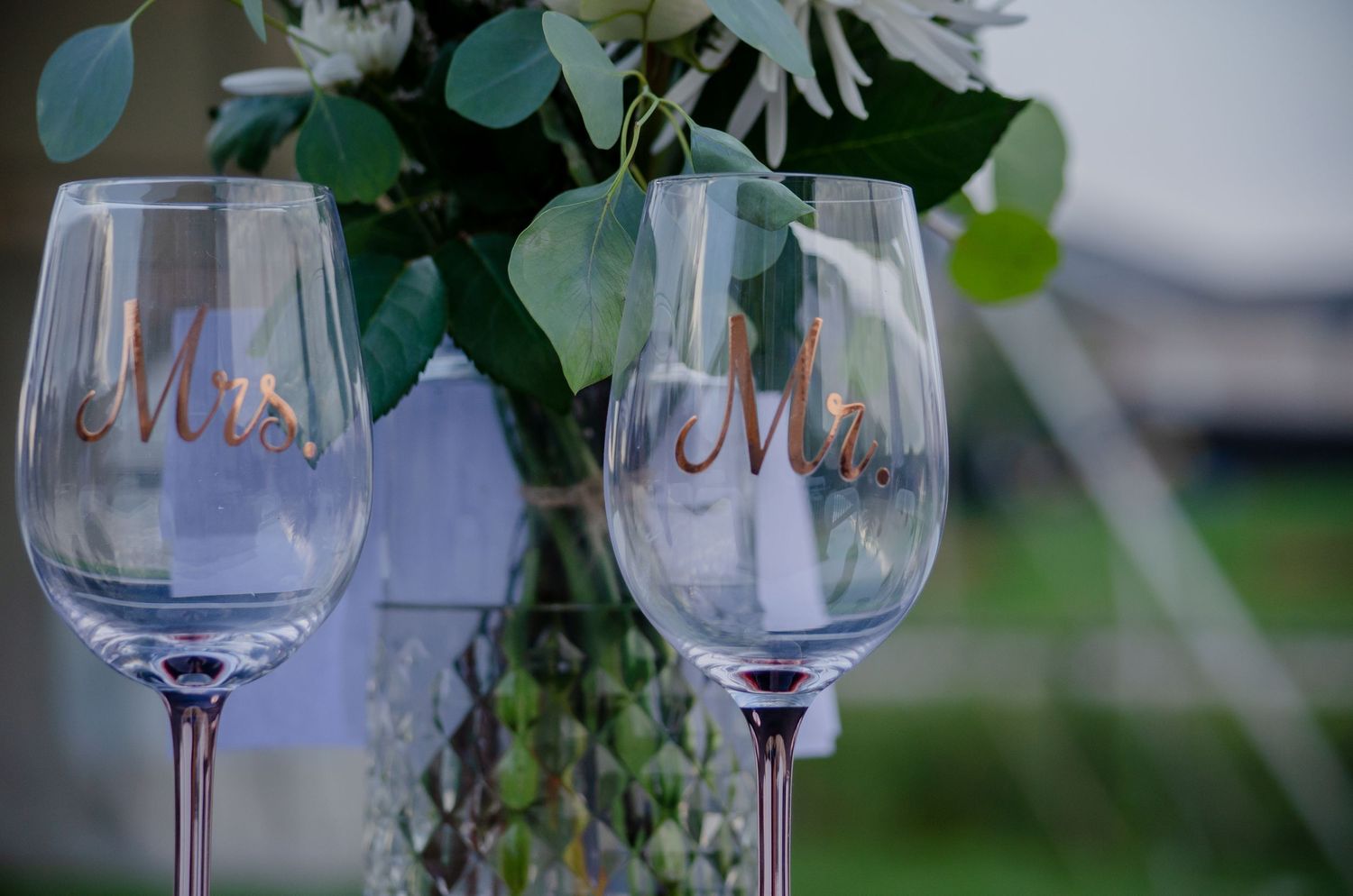 Mrs. and Mr. labeled wine glasses at a wedding reception with floral centerpiece in background.