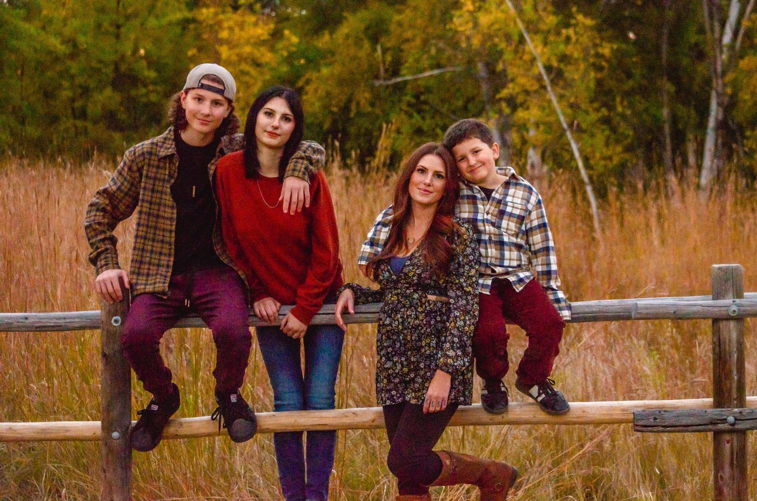 Family of four posing on a rustic wooden fence surrounded by autumn foliage in a scenic outdoor setting.