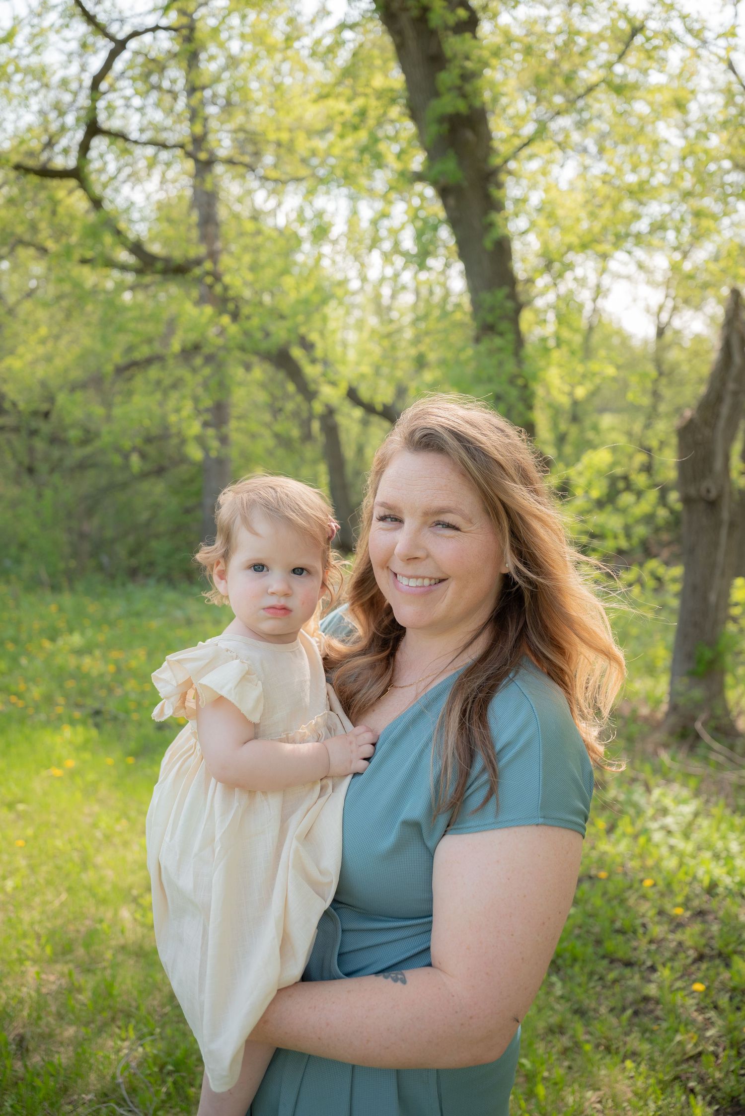 A smiling woman in a teal dress holds a toddler girl in a cream dress outdoors among green trees.