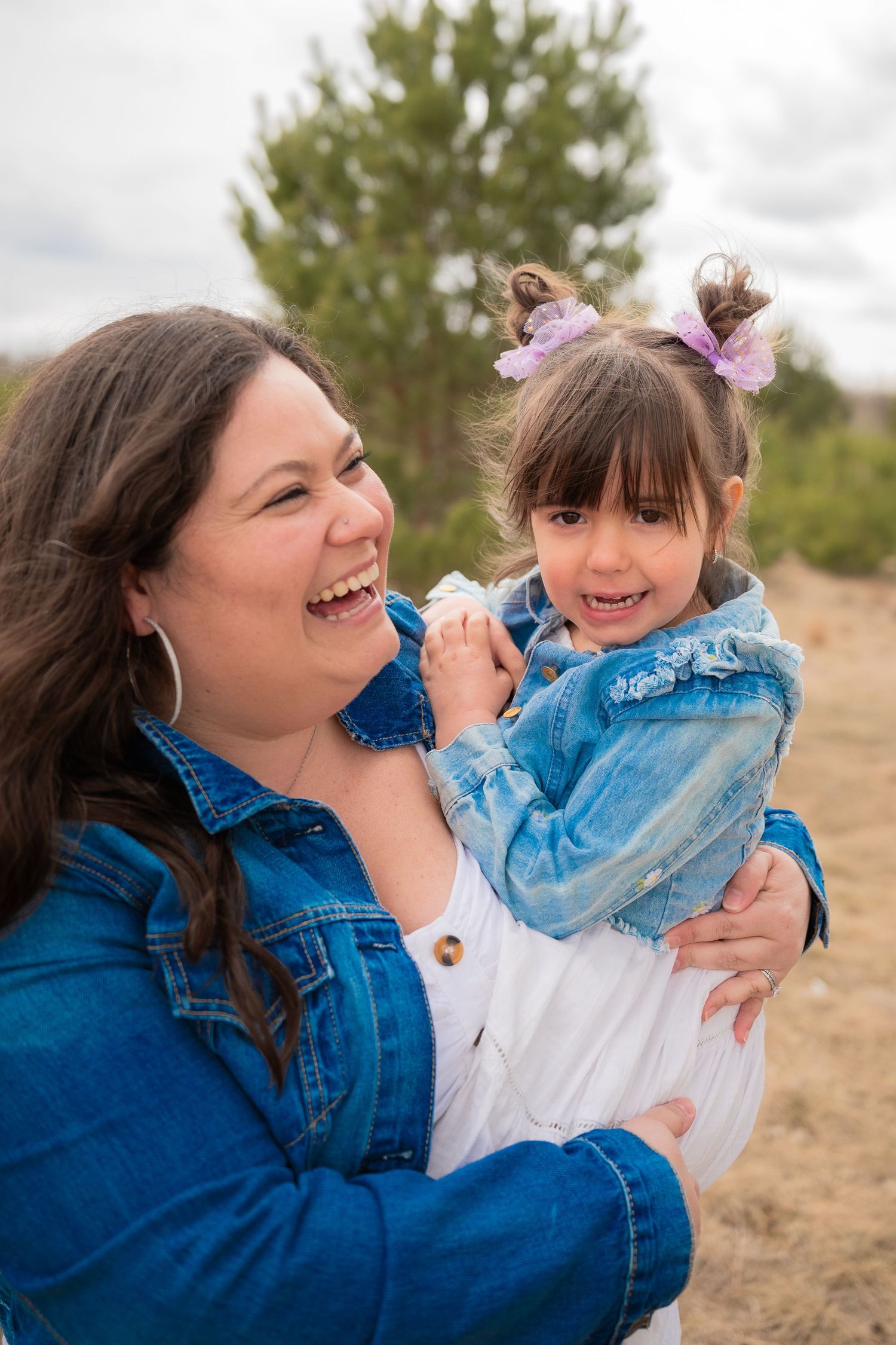 A laughing woman in a denim jacket holds a smiling little girl with a purple bow outdoors.