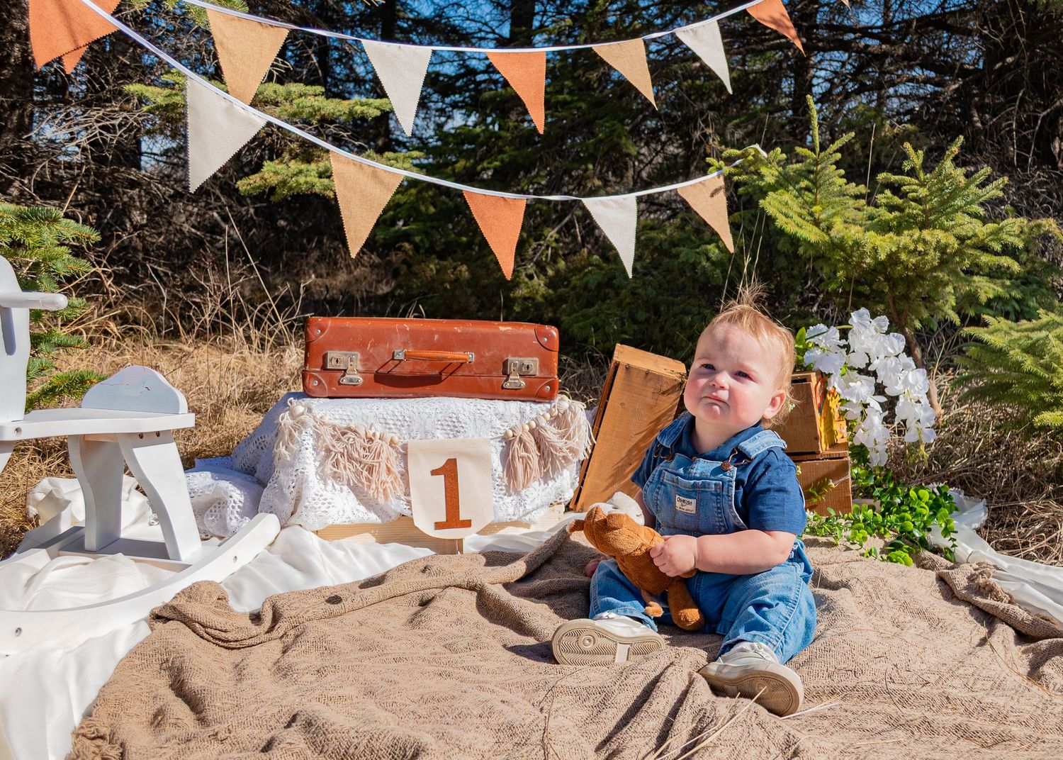 Baby boy in denim overalls sits on burlap at outdoor first birthday photo shoot with bunting and vintage suitcase.