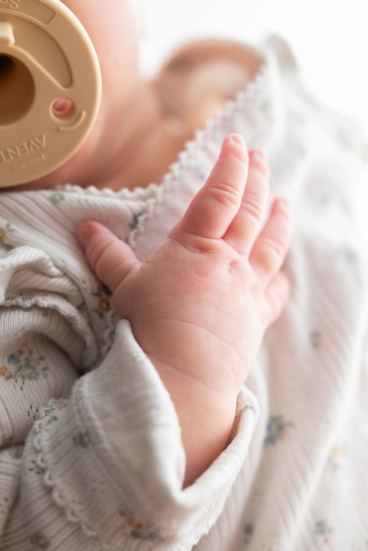 Close-up of a newborn baby's tiny hand resting on a floral onesie with a pacifier nearby.