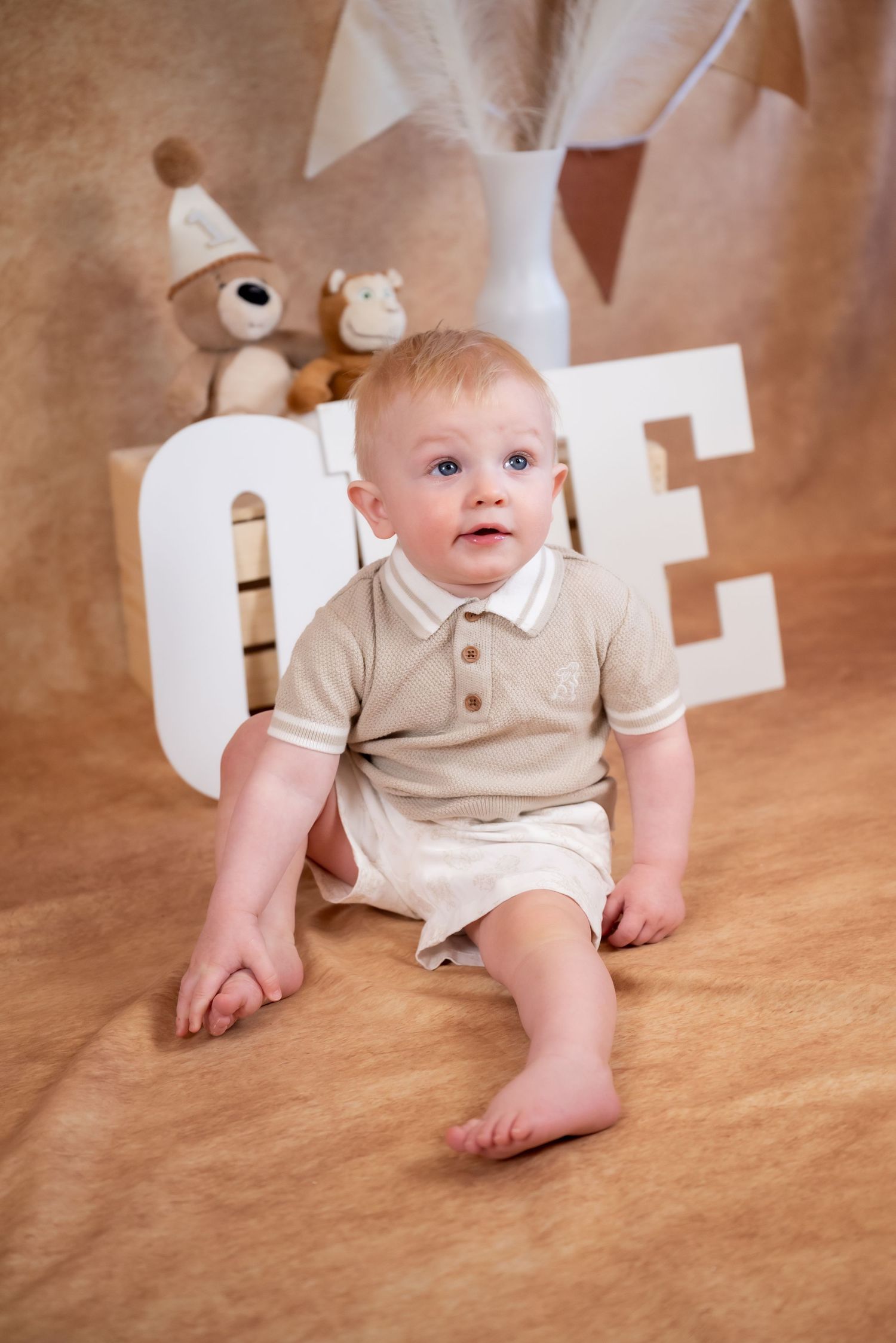 Adorable baby boy in beige romper sitting on floor with ONE letters and stuffed animals for first birthday photo.