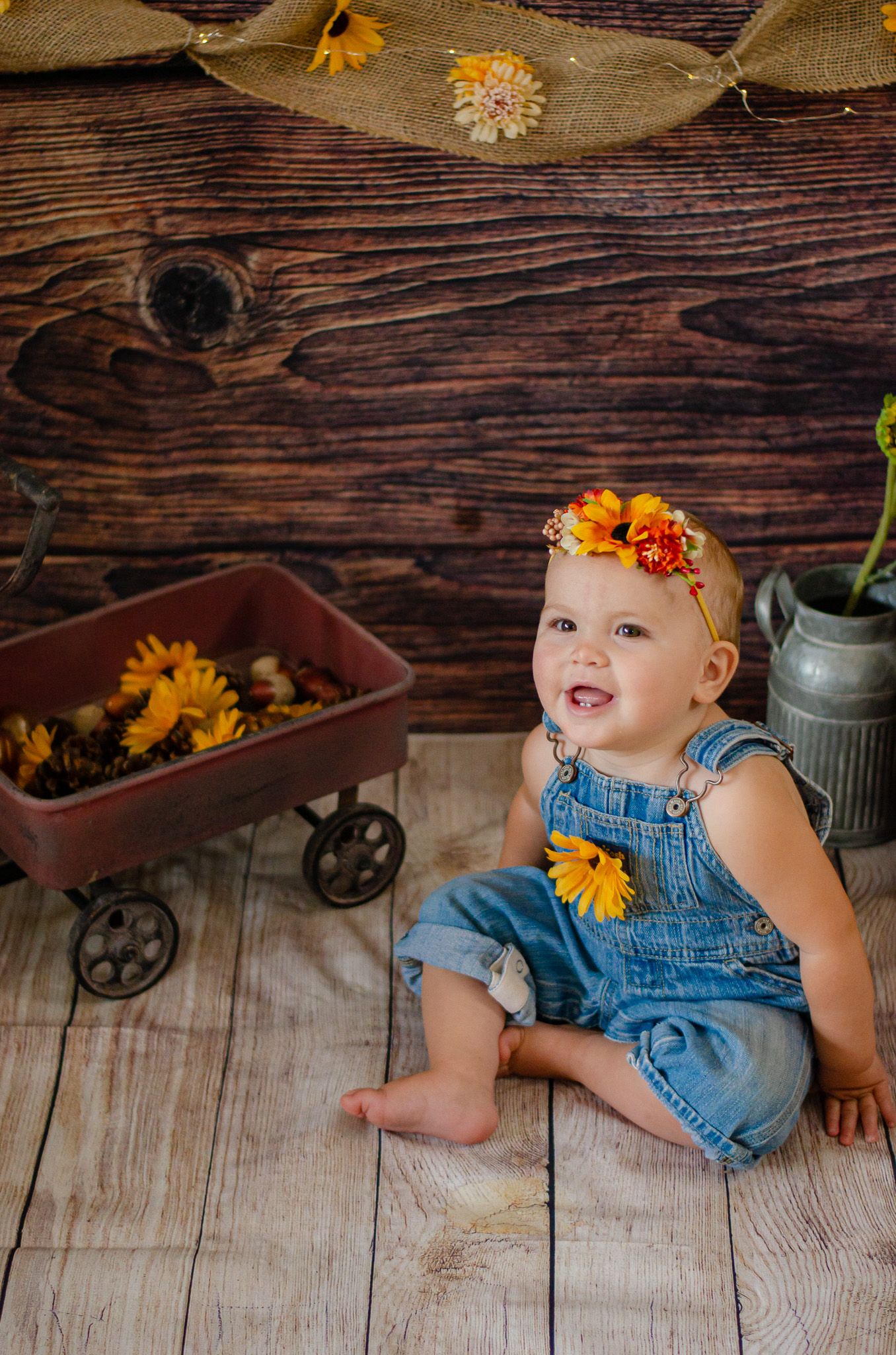 Smiling baby in denim overalls with sunflower headband sits on wood floor beside rustic wagon filled with sunflowers.