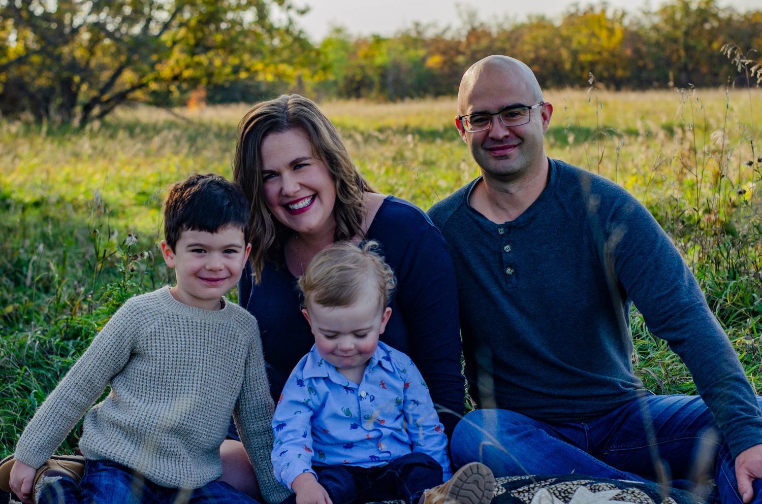 Family of four smiling outdoors in a sunny green field, parents with two young boys enjoying a warm autumn day.