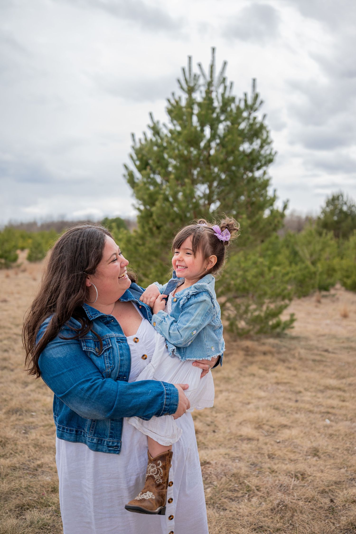 A mother holds her smiling toddler daughter outdoors near pine trees, both wearing white dresses and denim jackets.