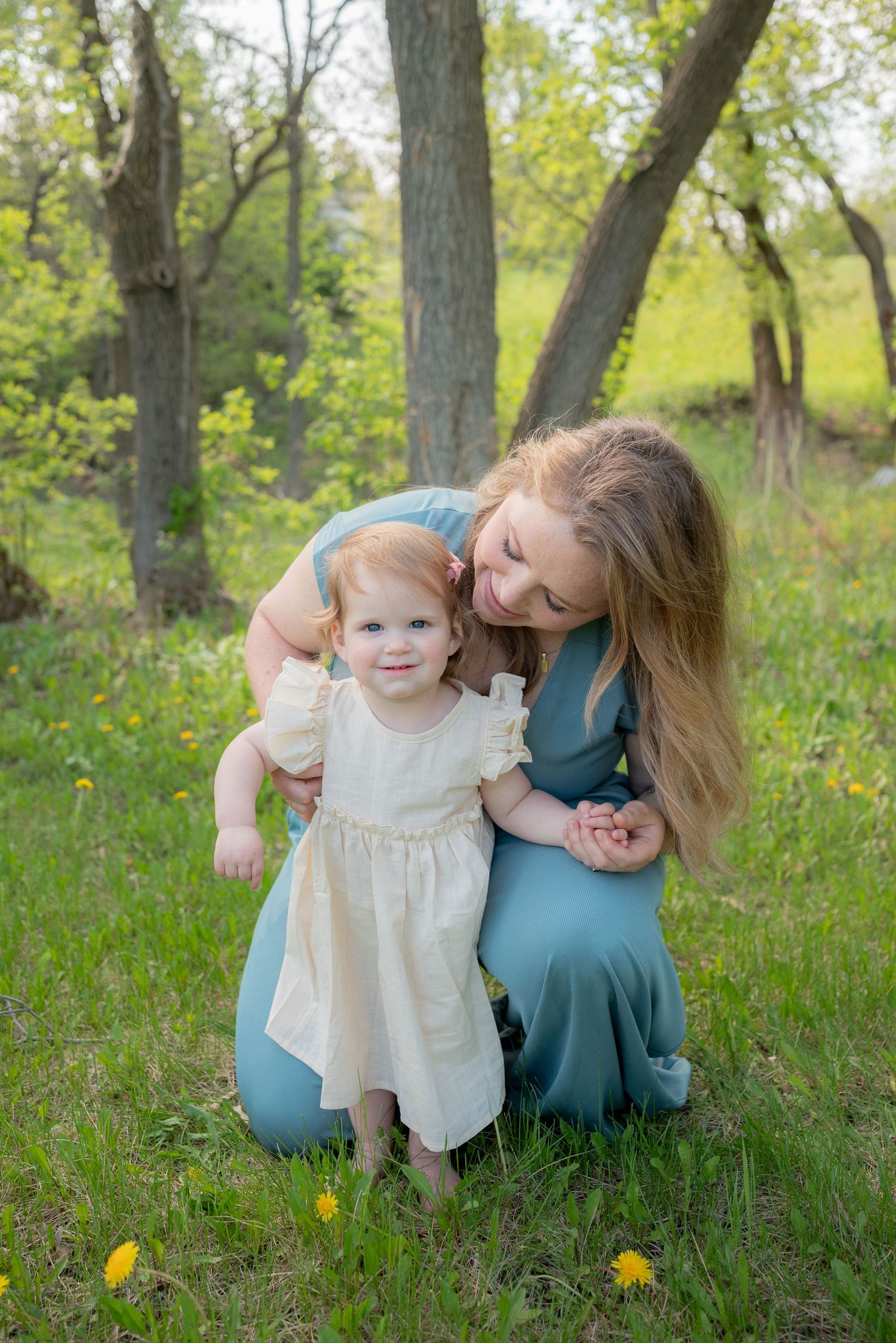 A young mother in a teal dress kneels beside her toddler girl in a white dress in a lush green outdoor setting.