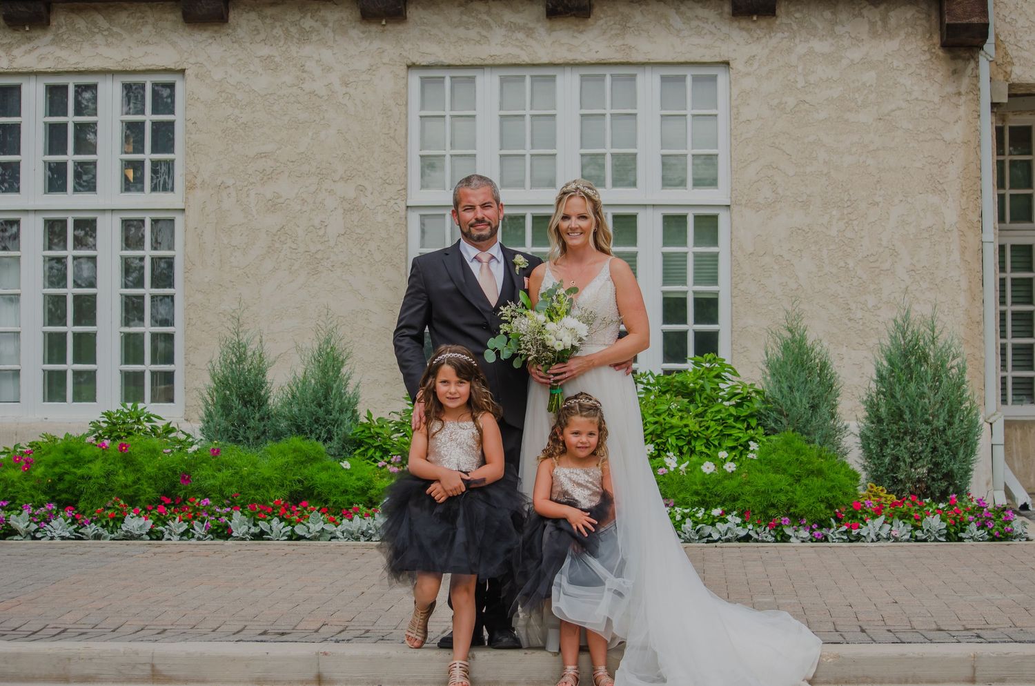 Bride and groom pose with two flower girls in tulle dresses outside an elegant venue with blooming gardens.