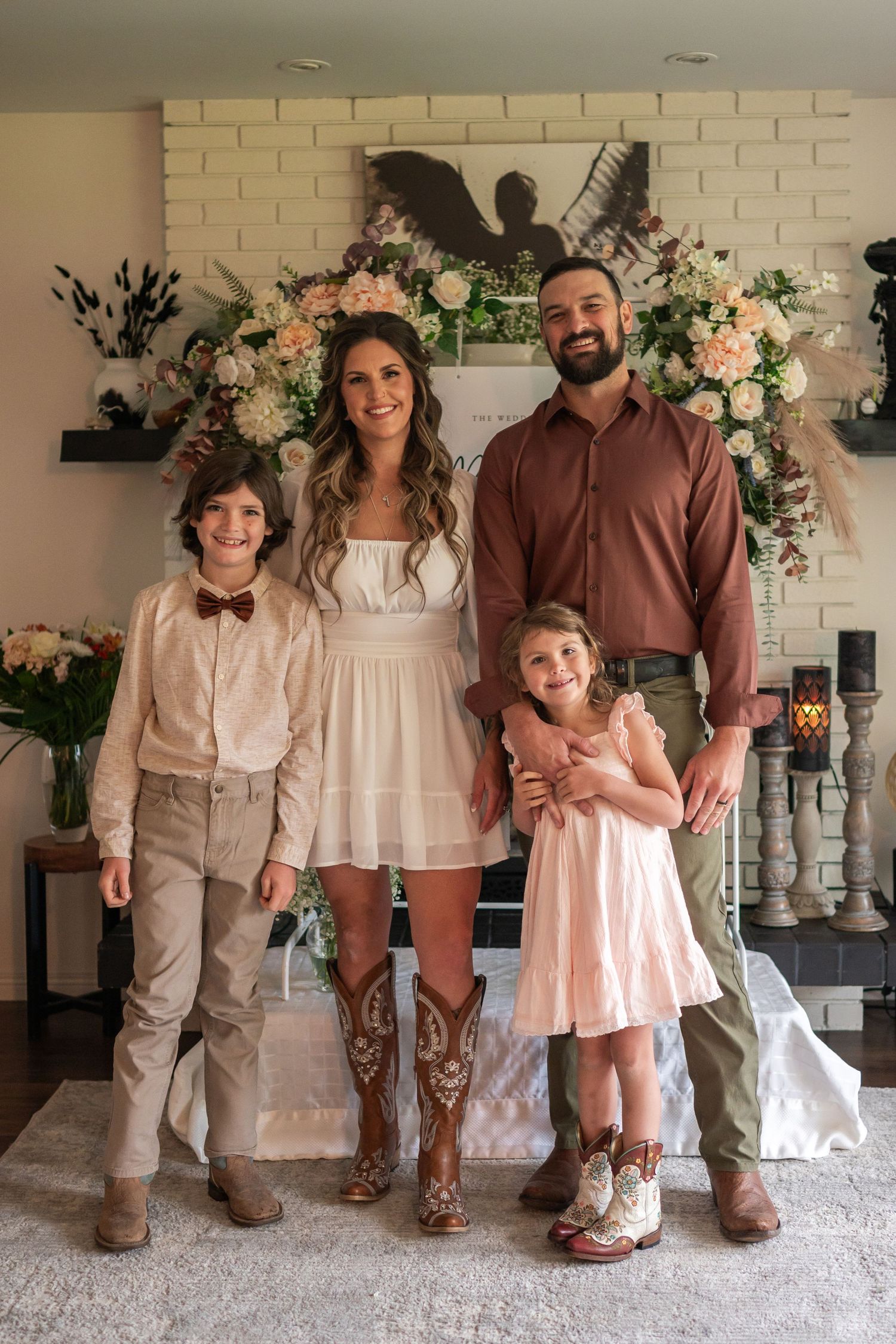 Family of four posing indoors with floral backdrop, wearing coordinated outfits including cowboy boots and bow tie.