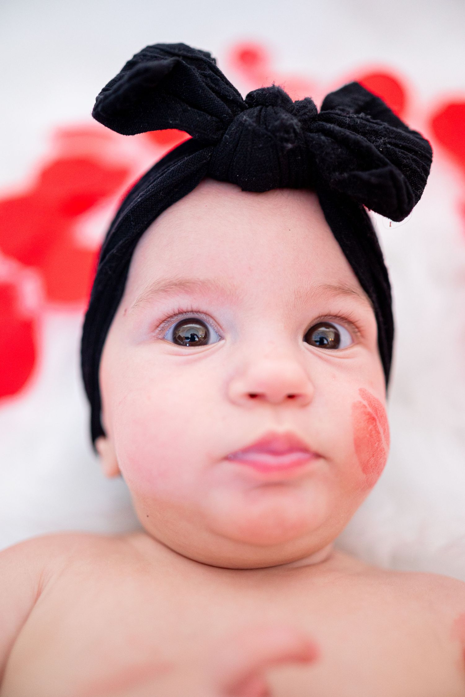 Adorable baby with a black bow headband and a red lipstick kiss mark on cheek, looking up with big brown eyes.