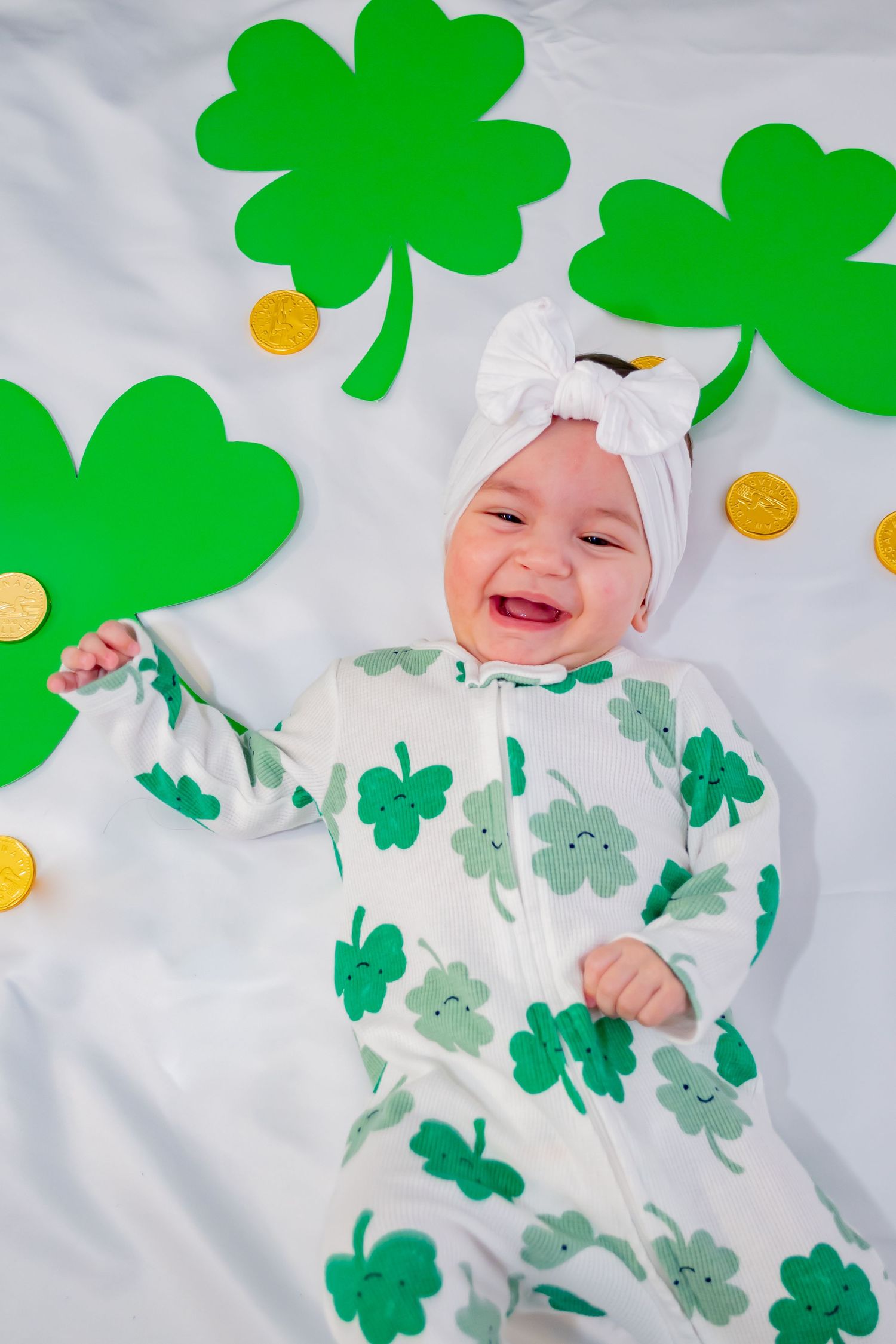 Laughing baby in shamrock onesie and white bow turban laying among green clover decorations and gold coins for St. Patricks Day.
