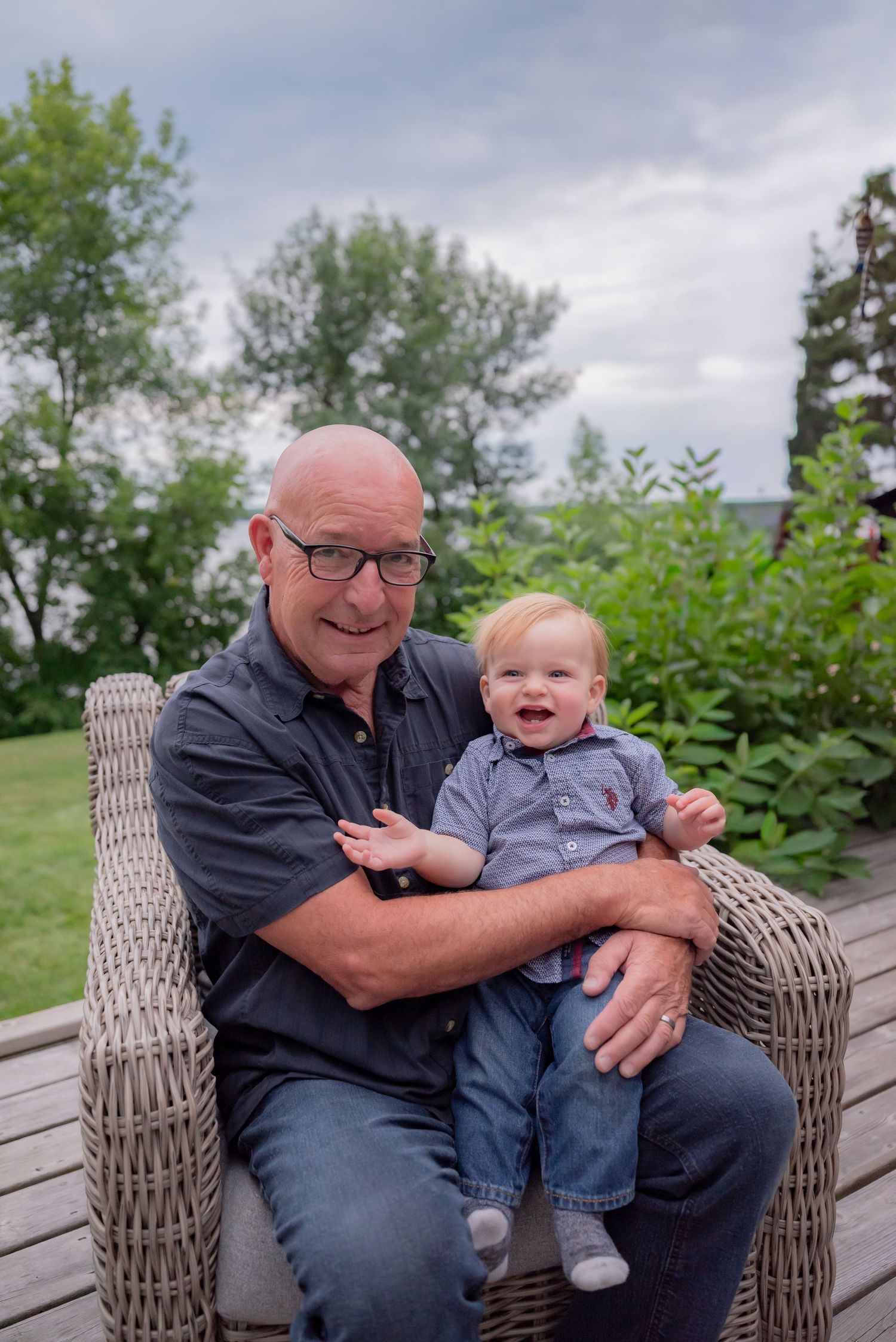 Bald man with glasses smiling while holding a happy baby on a wicker chair outdoors with green trees.