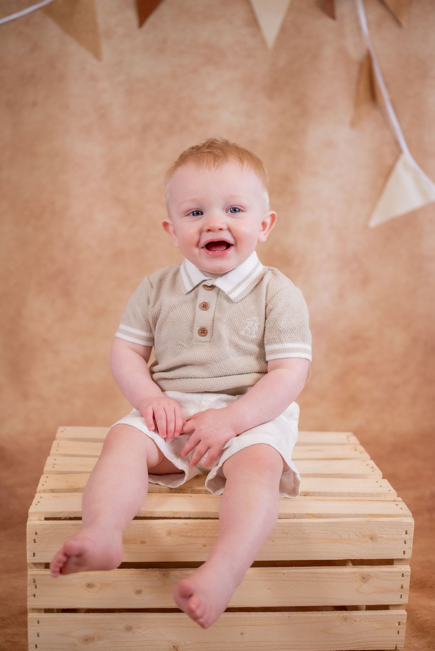 Happy baby boy in beige outfit sitting on wooden crate, smiling, with bunting backdrop in professional photo studio.