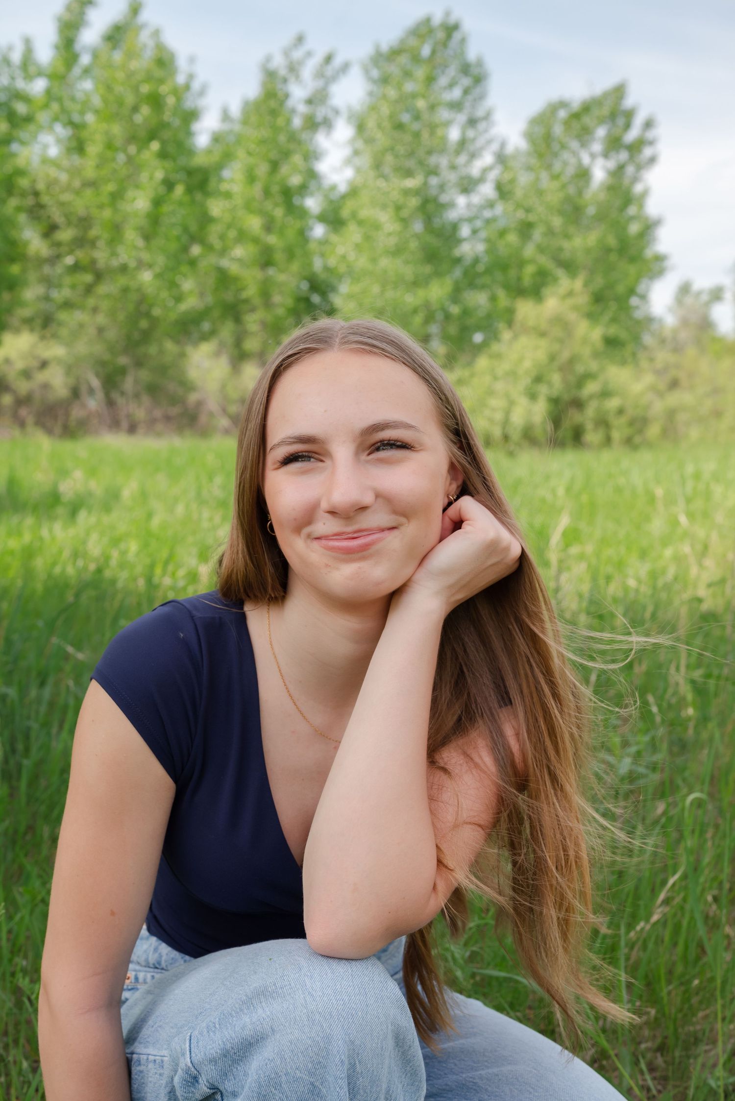 Smiling young woman with long hair sitting in a green field, wearing a navy top and jeans.