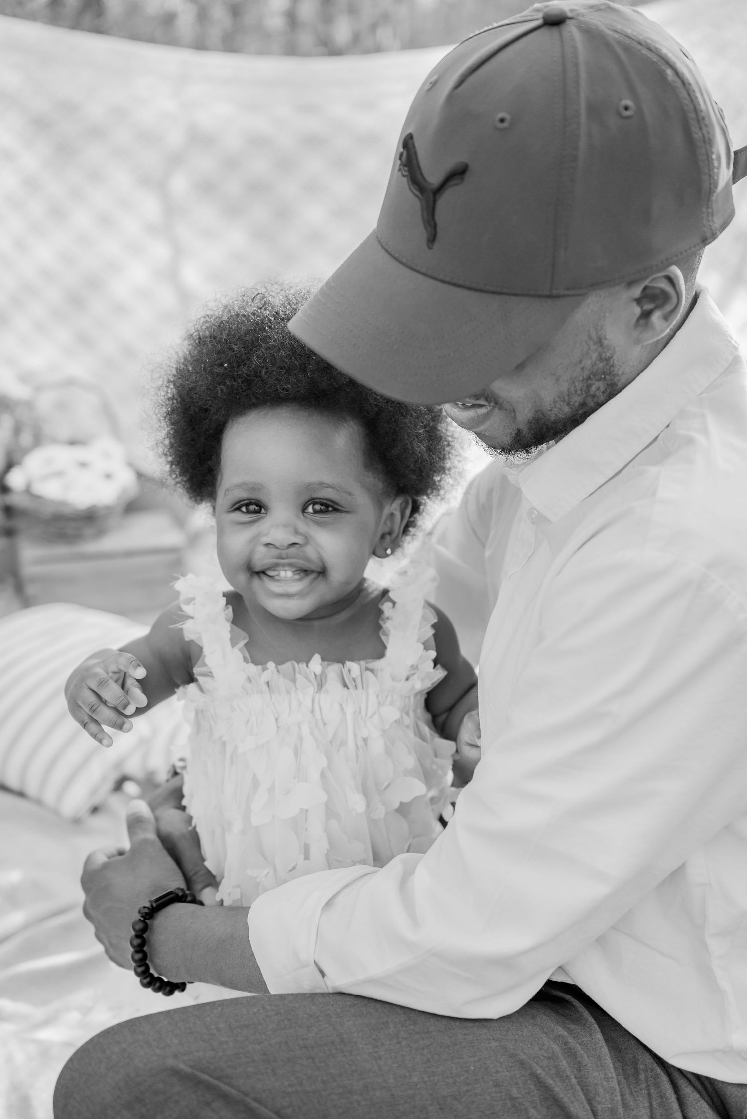 Black and white photo of a smiling toddler girl held by a man wearing a Puma cap outdoors.
