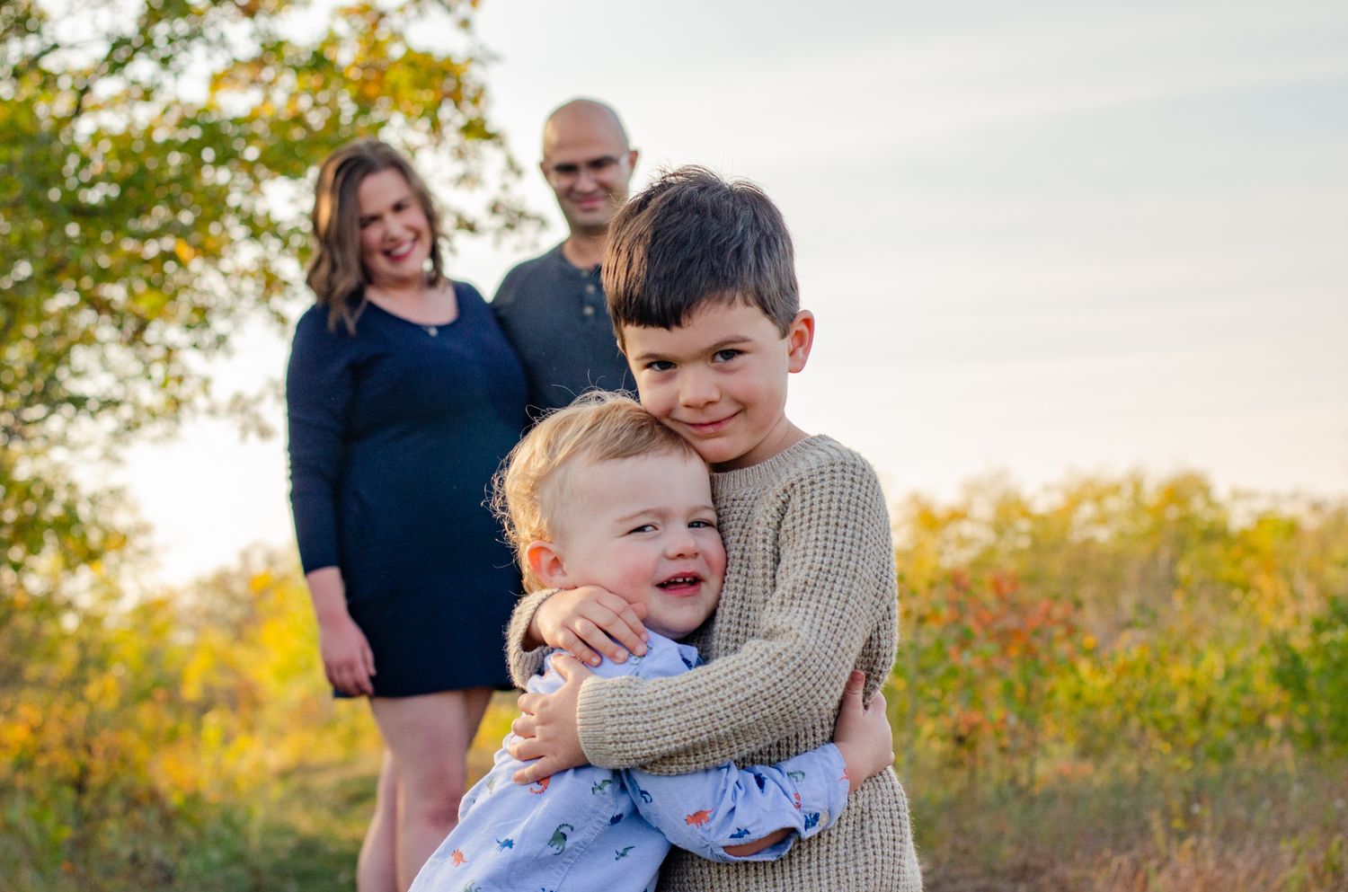 Two brothers share a warm hug outdoors in autumn, with their parents smiling in the background.