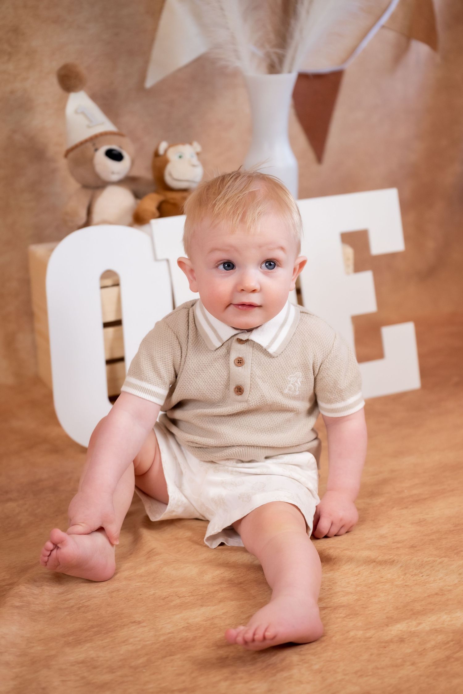 Adorable baby boy in beige outfit sitting in front of ONE letters and teddy bears for first birthday photo.