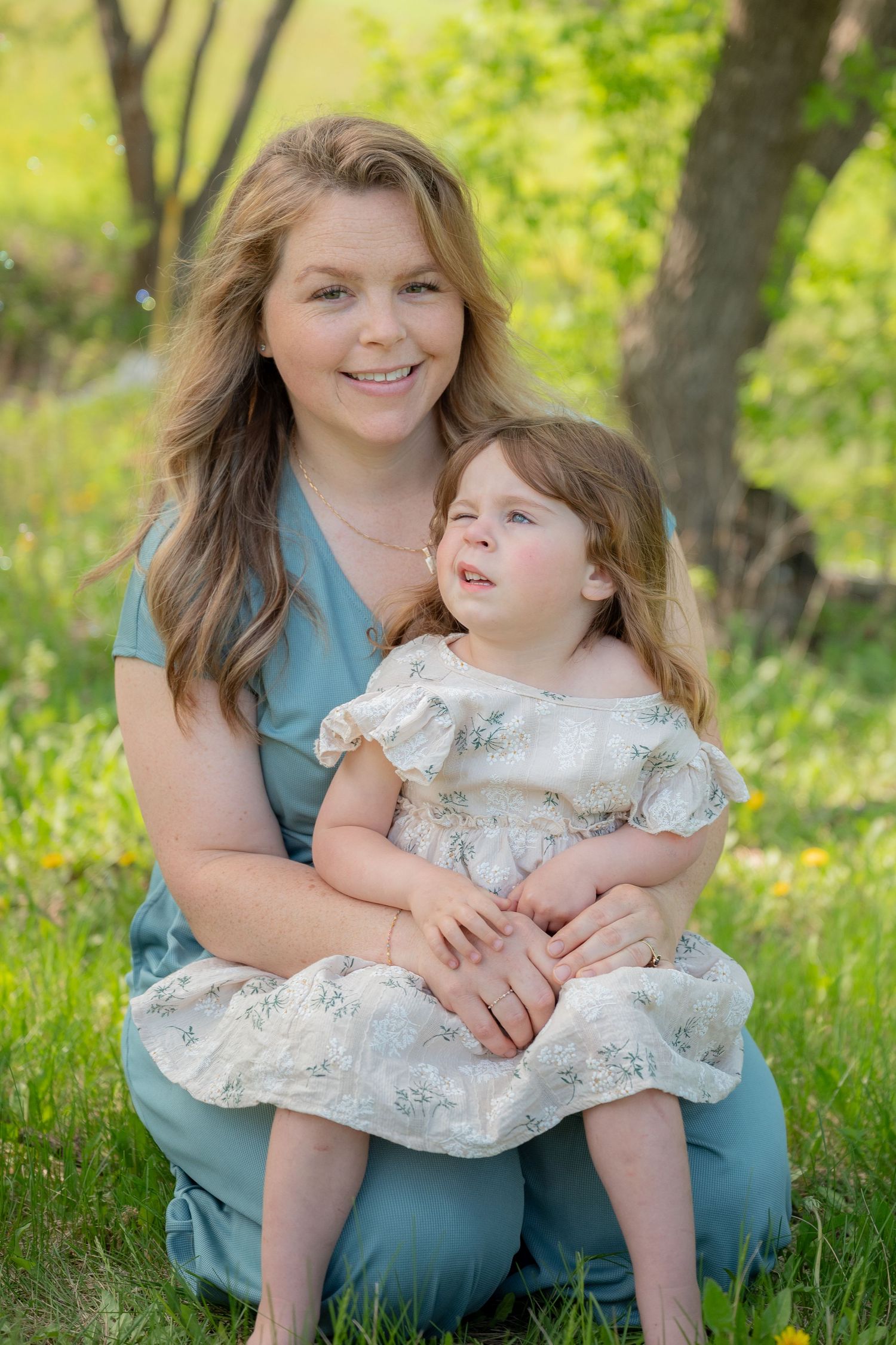 A smiling woman in a teal dress holds a young girl in a floral dress outdoors in a lush green park setting.