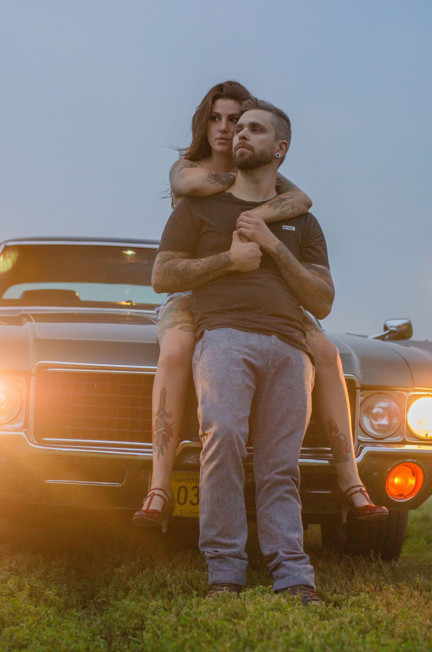 Man holding woman in his arms in front of a classic muscle car at dusk with warm glowing headlights.