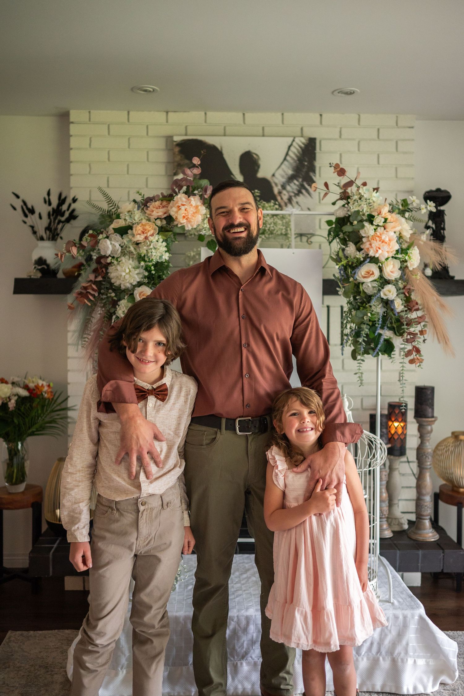 Man with two children smiling in front of a floral decorated fireplace mantel indoors.
