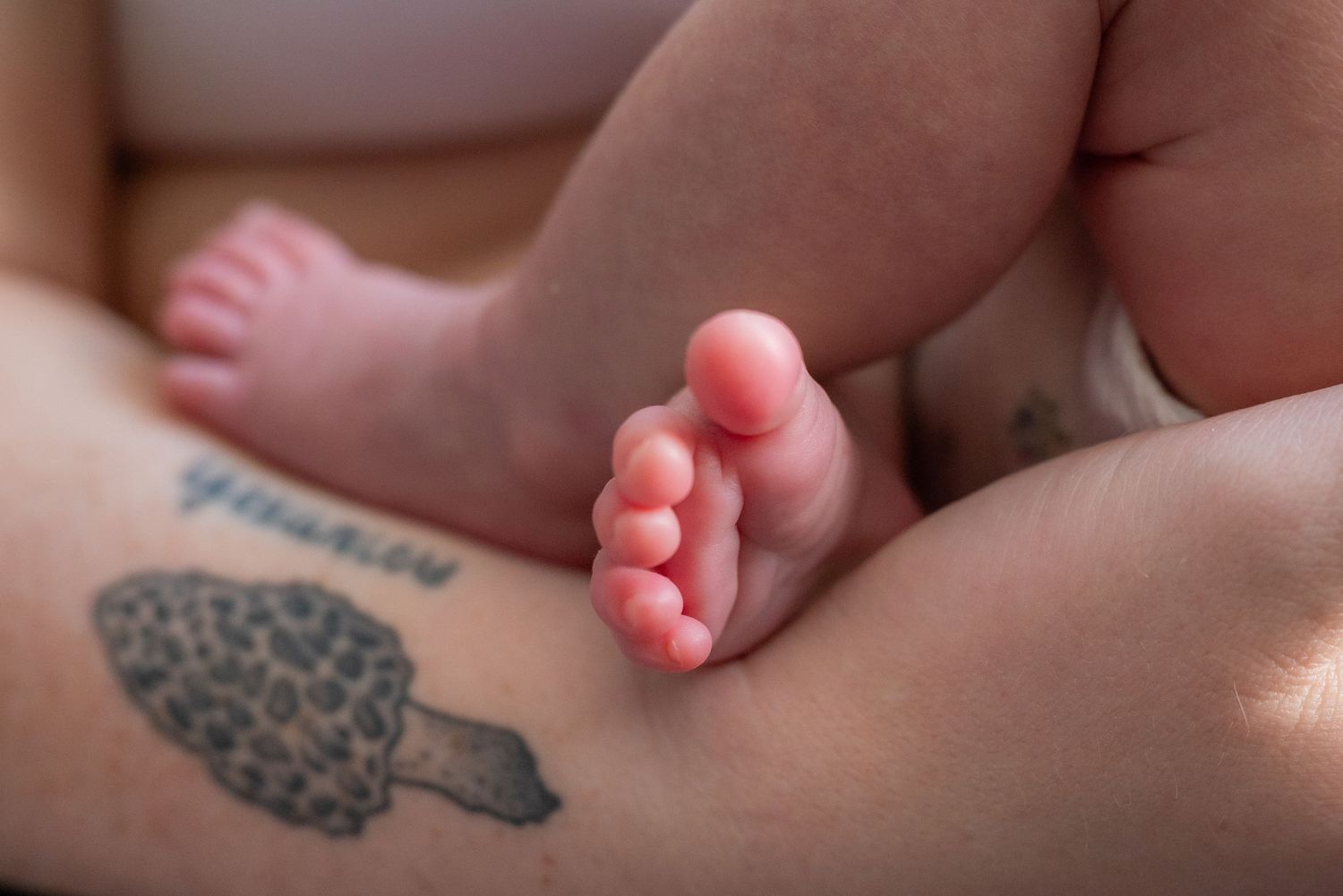 Close-up of newborn baby feet resting on mother's leg with a tattoo, capturing a tender postpartum moment.