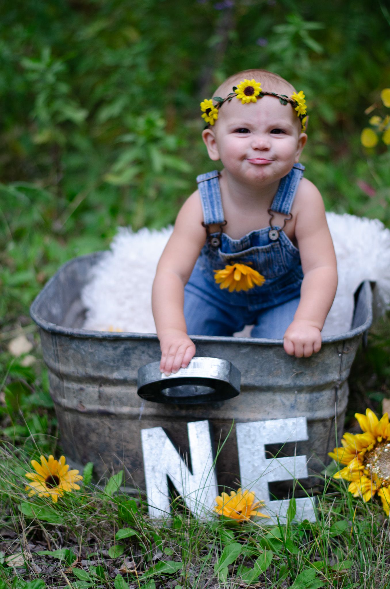 Baby in denim overalls and sunflower crown sits in vintage metal tub outdoors, surrounded by sunflowers and ONE letters.