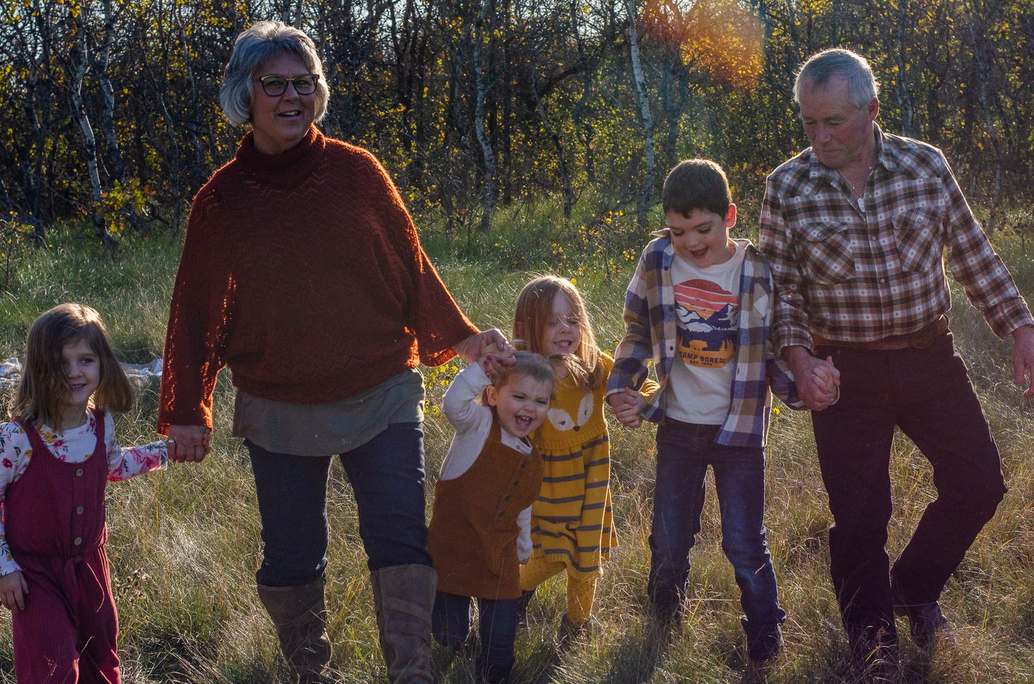 Two grandparents walk outdoors in a field with three young children during autumn, holding hands and smiling.
