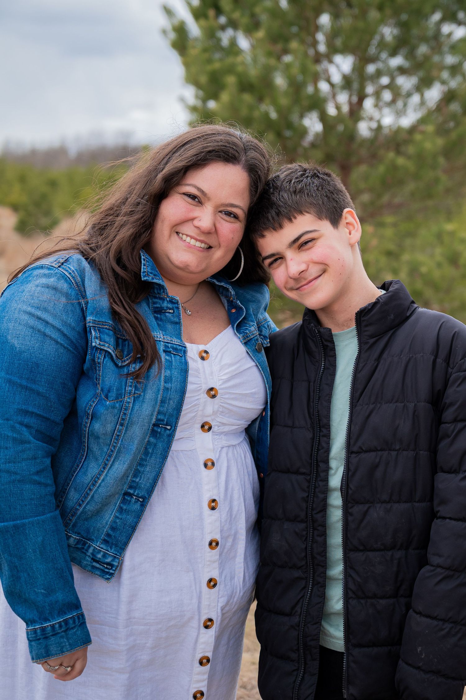 A smiling woman in a denim jacket and white dress poses with a teenage boy outdoors near pine trees.