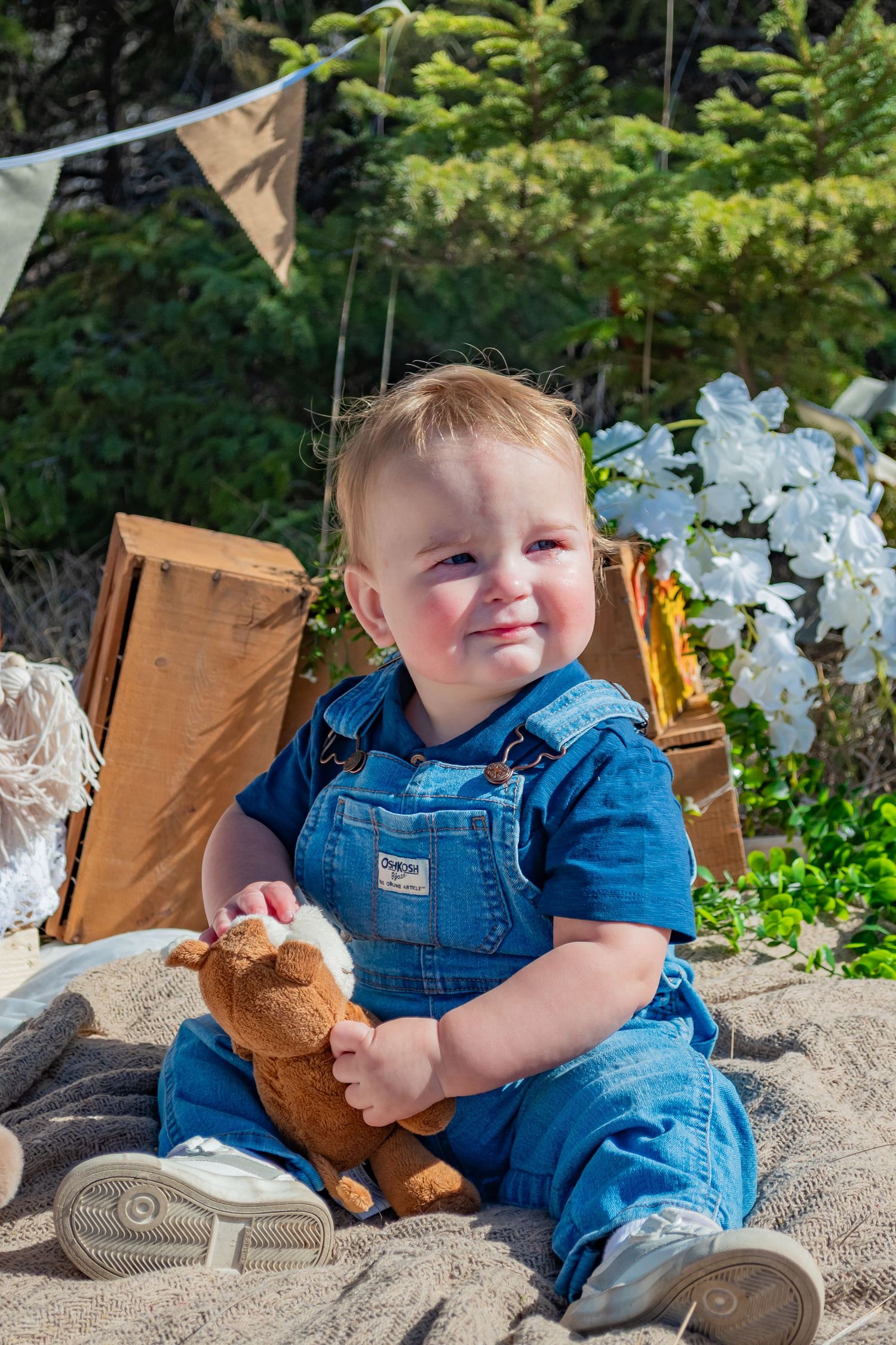 Smiling baby in denim overalls sitting outdoors holding a stuffed animal toy with vintage decor and greenery behind.
