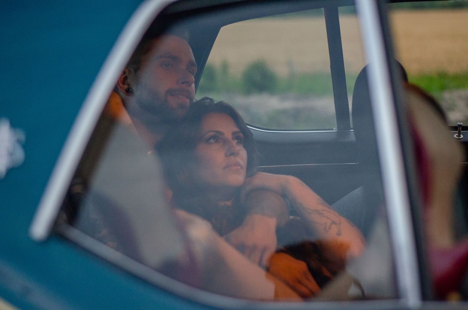 A couple sitting together in a vintage blue car, the woman leaning on the man's shoulder, looking contemplative.