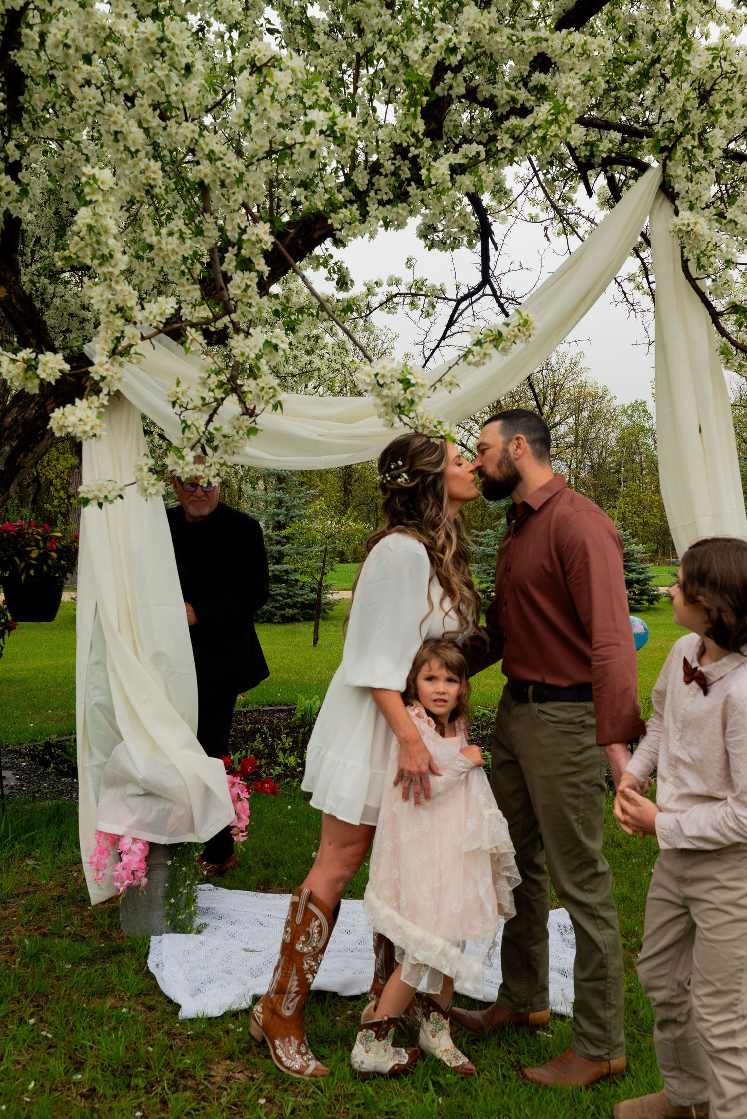 Couple shares first kiss at outdoor wedding ceremony under white floral arch with flower girl and guests nearby.